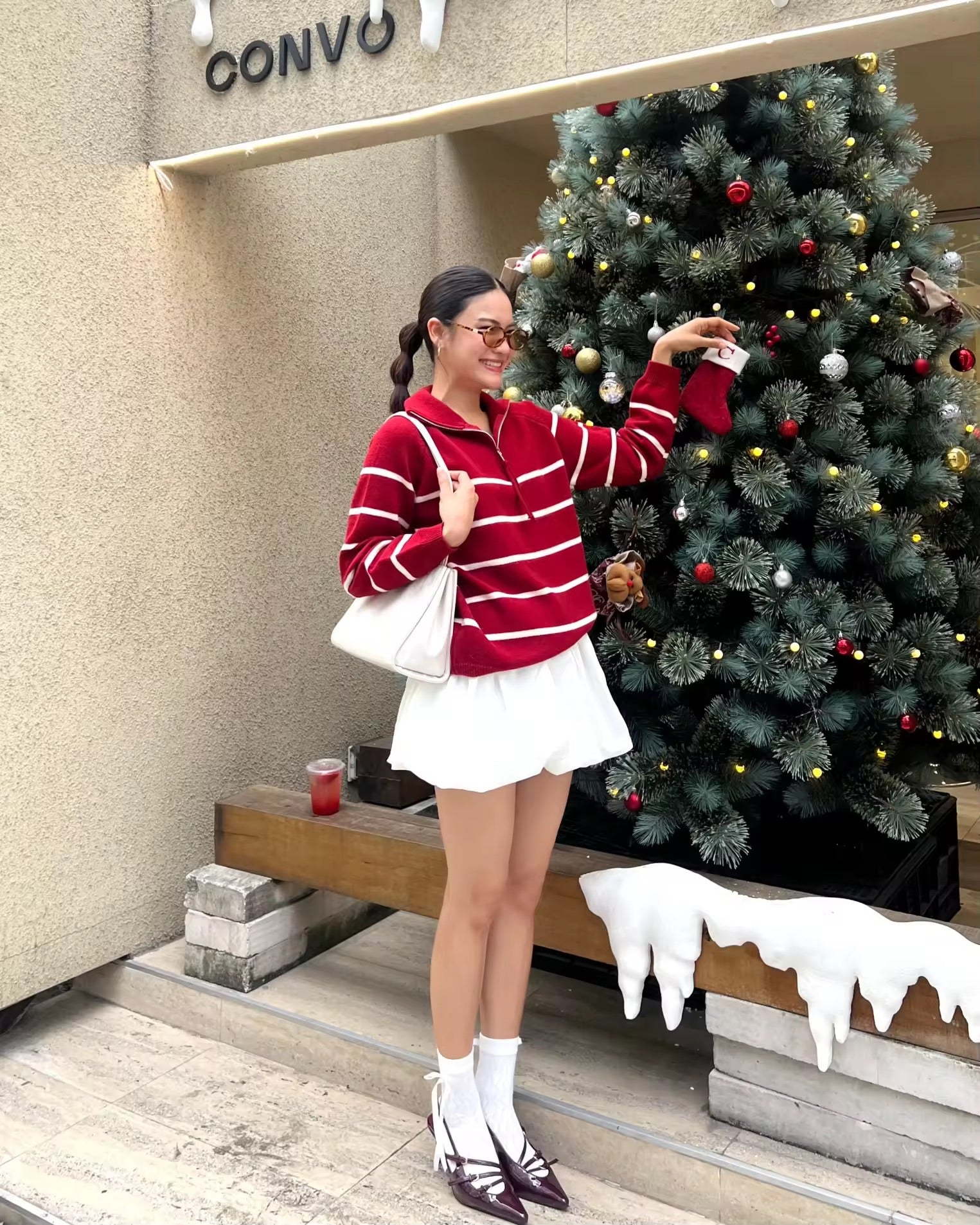 Woman in a red and white striped sweater and white skirt standing next to a Christmas tree in front of a CONVO store.