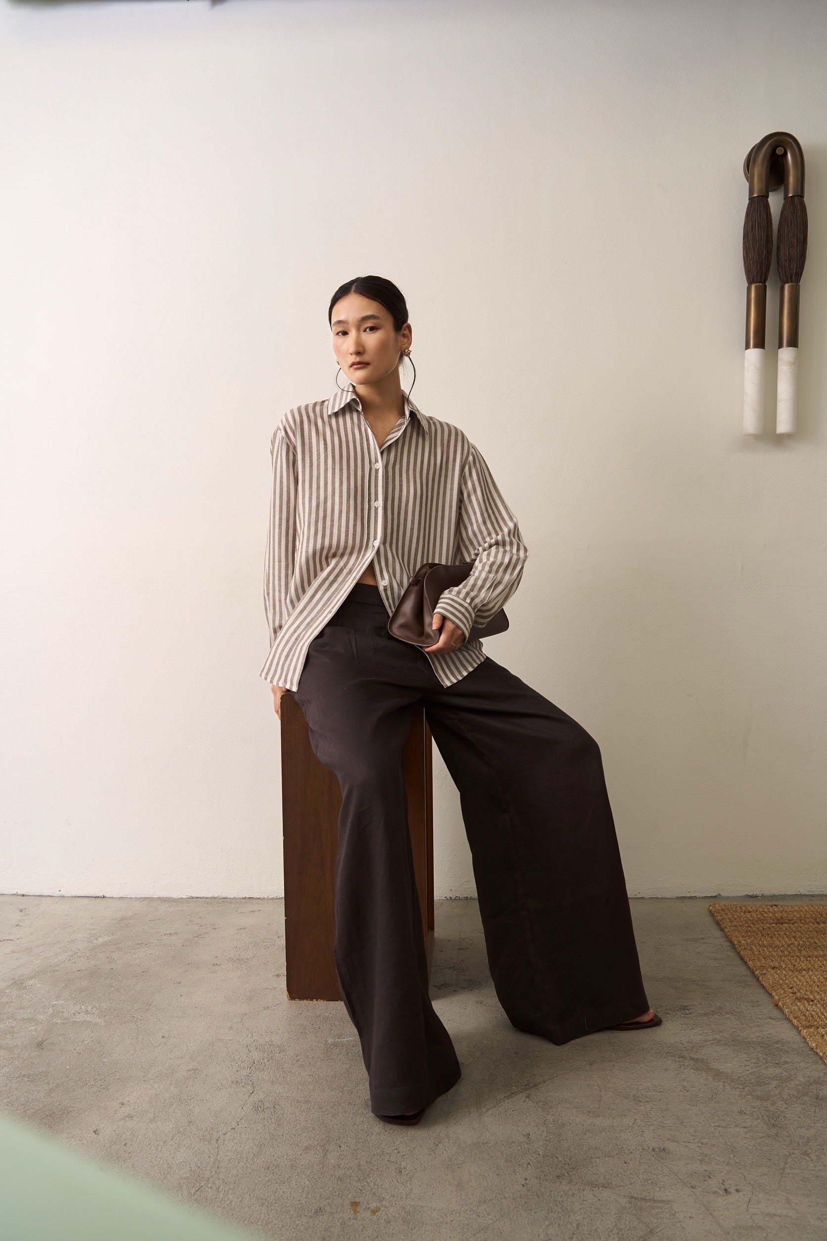 Woman sitting on a wooden stool wearing a brown striped linen shirt and brown pants in a plain background setting.