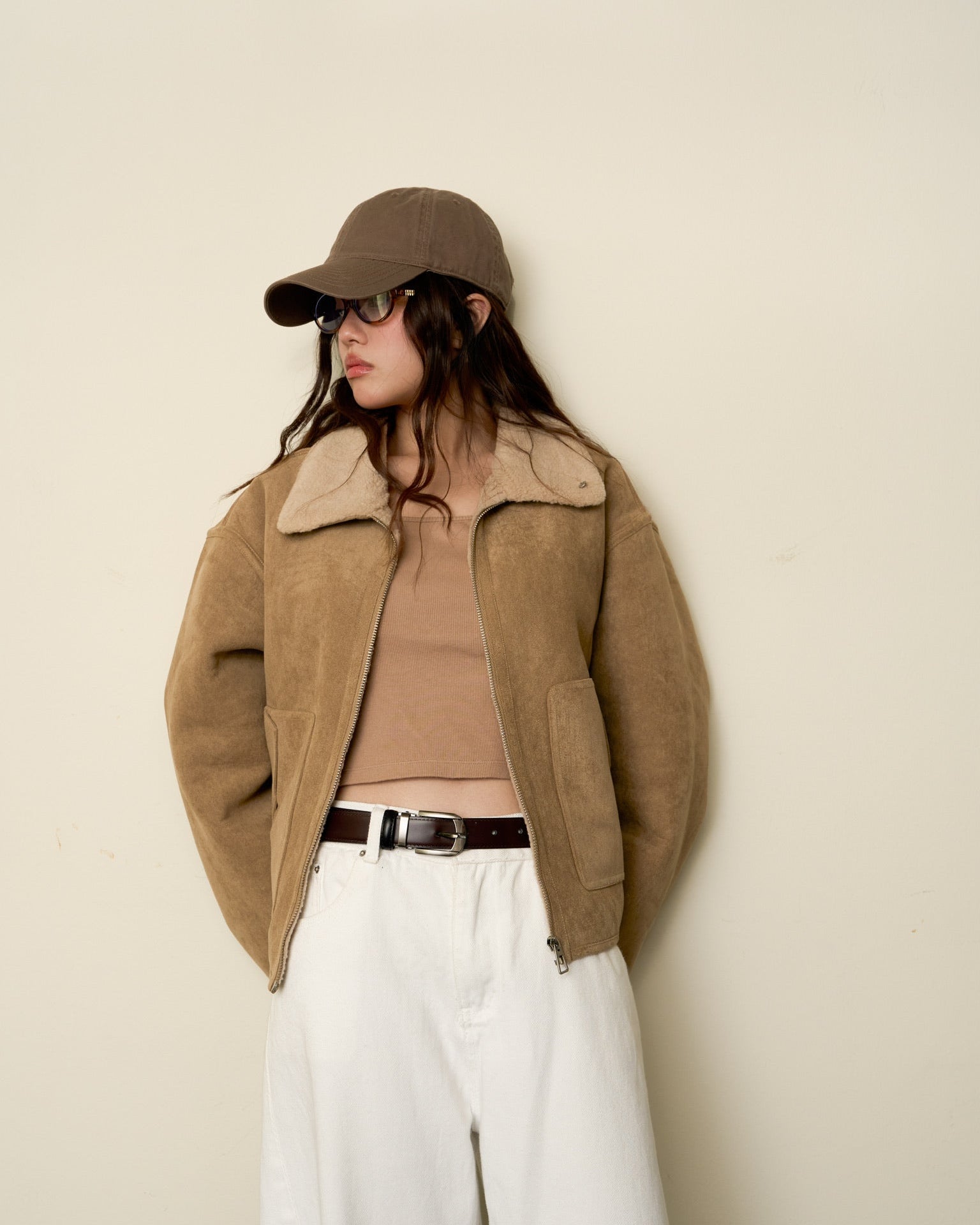 woman wearing a beige jacket, brown cap, and white pants against a plain background
