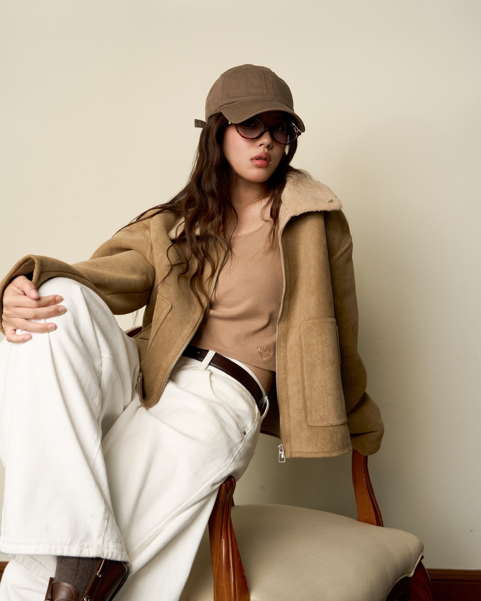 woman wearing a brown coat, beige sweater, and white pants sitting on a chair.