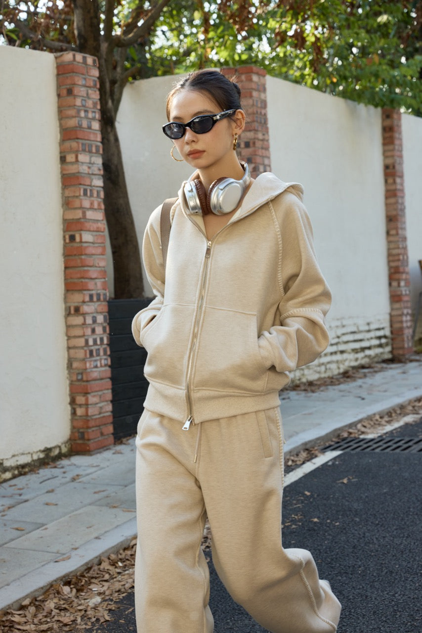 woman wearing a beige tracksuit with sunglasses and headphones, standing on a sidewalk.
