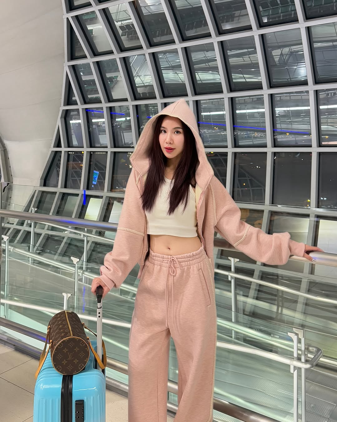 A woman stands in an airport terminal wearing a light pink hooded sweatshirt and matching sweatpants over a beige crop top, holding a light blue suitcase with a brown monogram bag hanging from the handle.