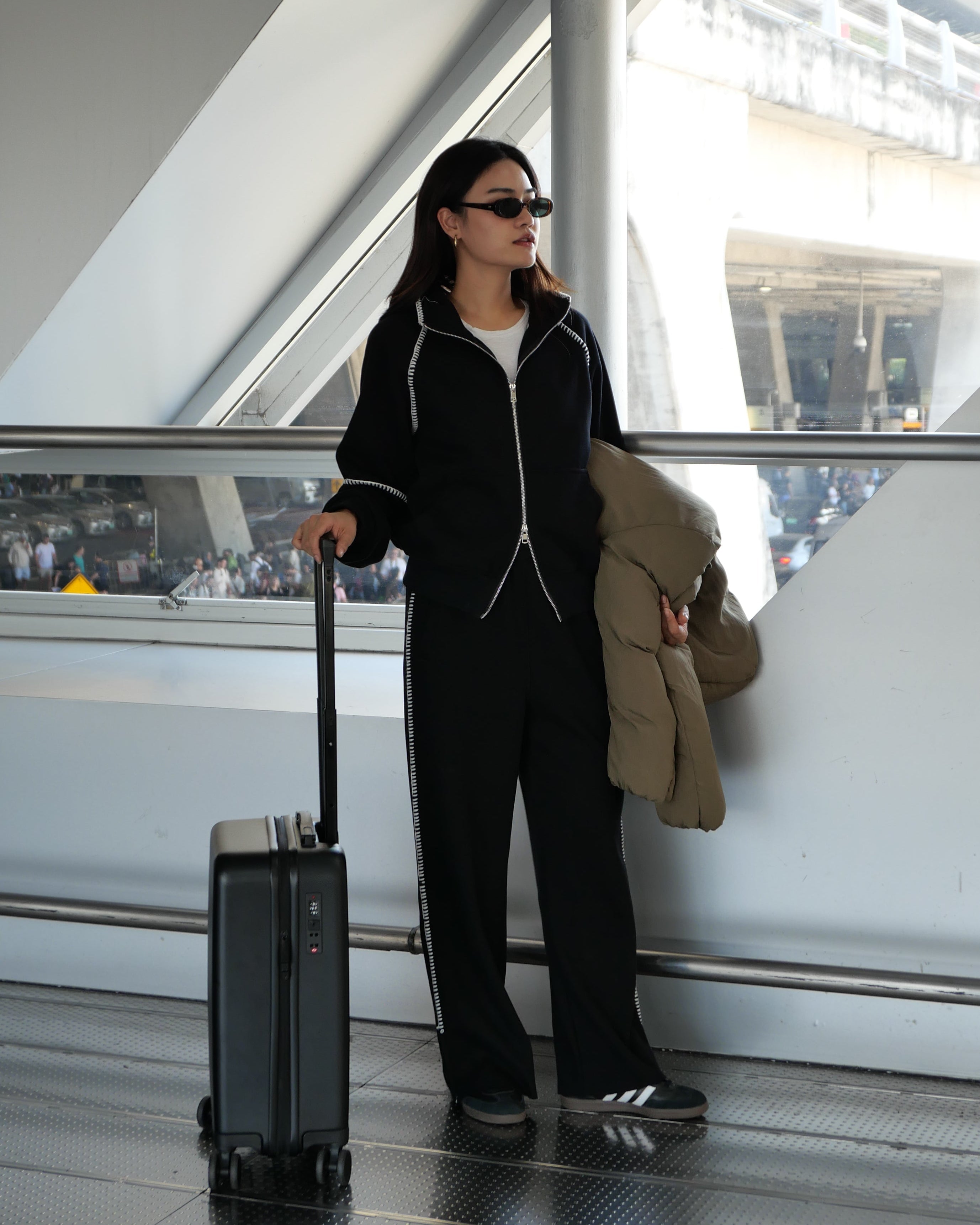 A woman in a black zip-up tracksuit and sunglasses stands in a modern indoor transit space, holding a beige coat and standing next to a black rolling suitcase.
