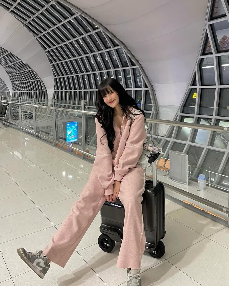 A woman posing in a modern airport terminal wearing a light pink two-piece lounge set, sitting on a black suitcase.