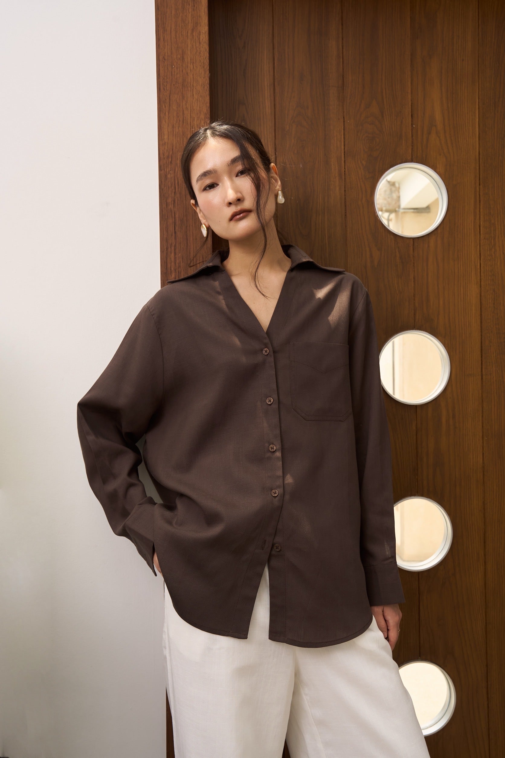 Woman wearing a brown linen shirt and white linen pants standing in front of a wooden door with circular lights.