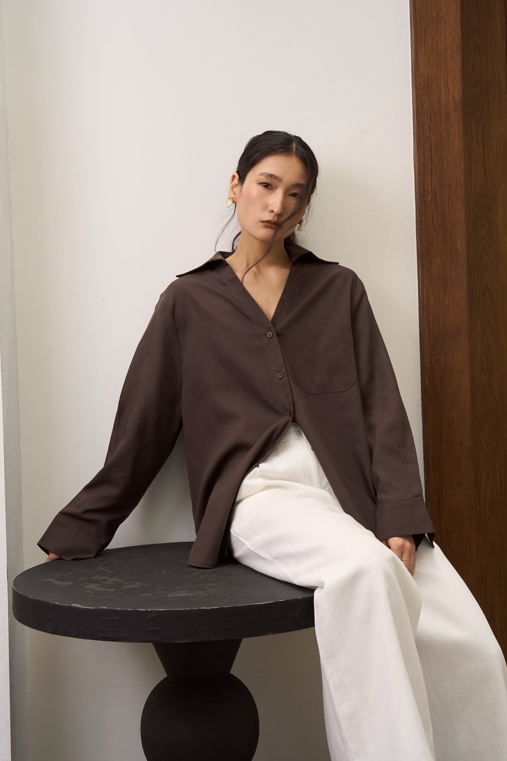 Woman in a brown v collar shirt and white linen pants sitting on a black stool against a plain wall.