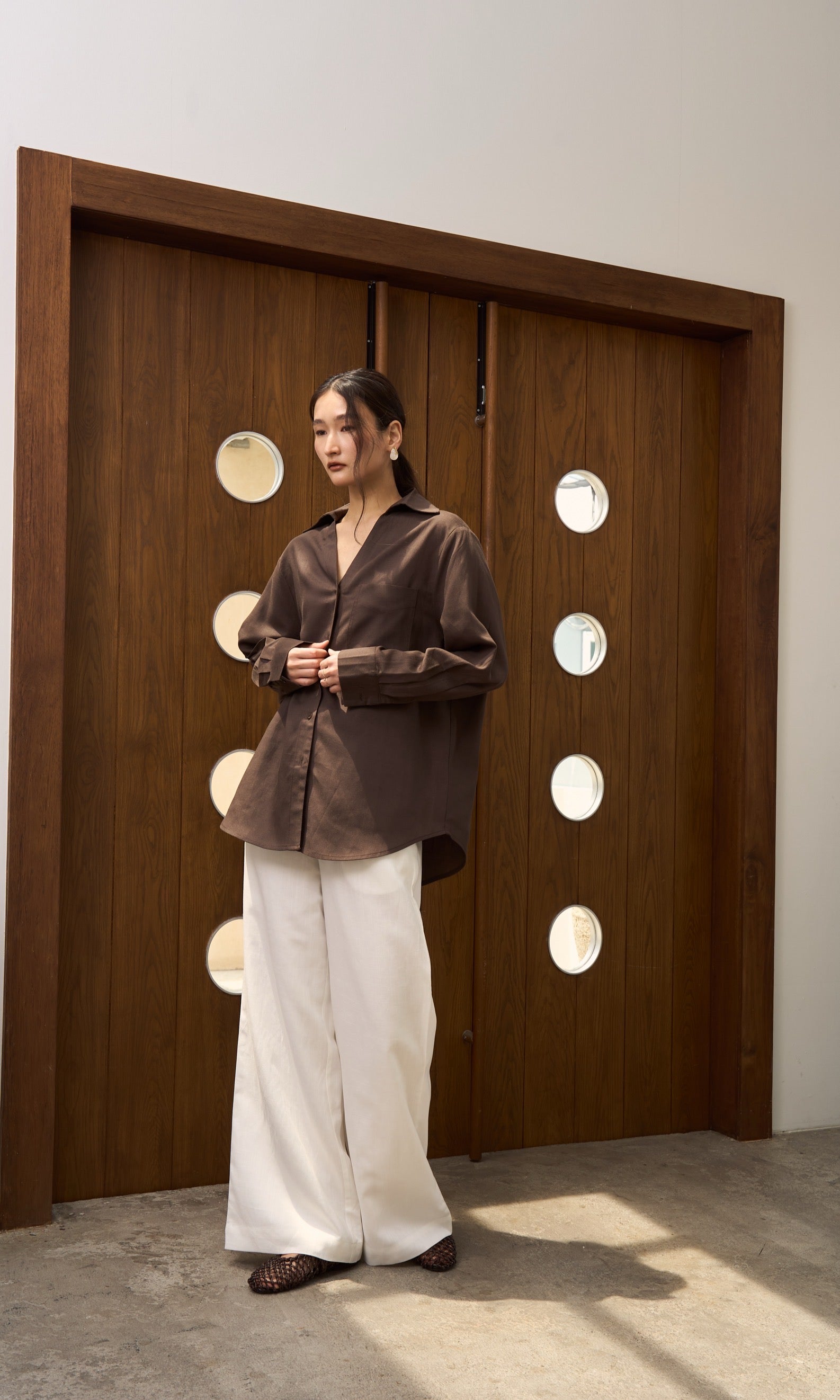 Woman in a brown v collar linen shirt and white linen pants standing in front of a wooden panel with circular lights.