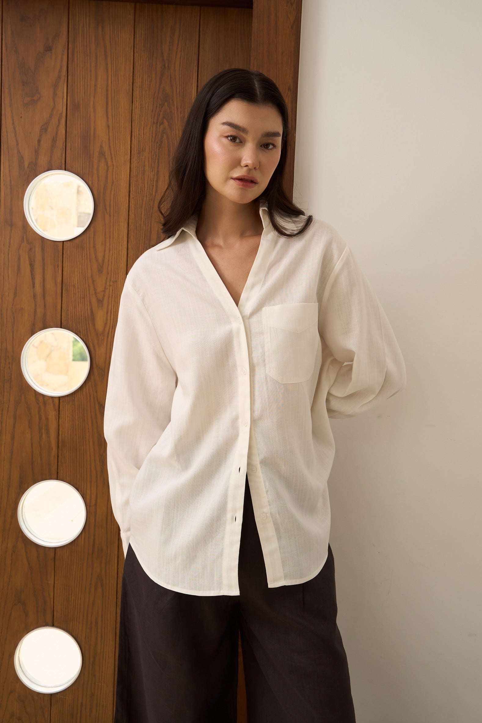 Woman wearing a white linen shirt showing v-collar and dark brown pants standing against a wooden wall with circular light fixtures.