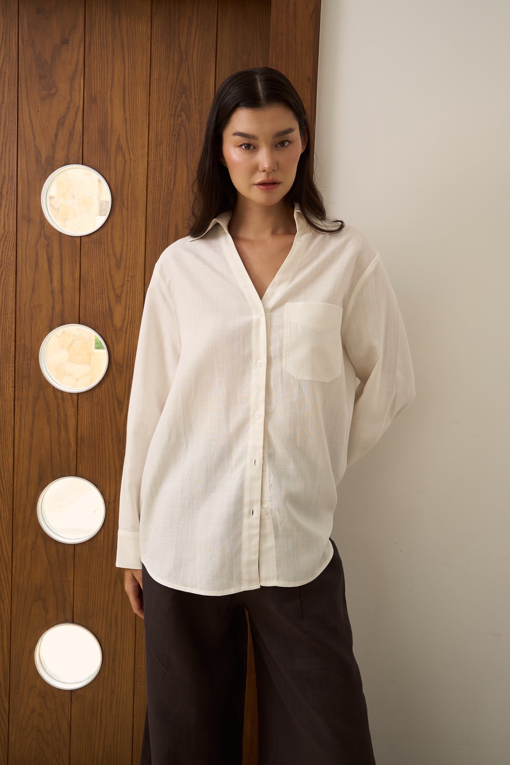 Woman wearing a white v-collar linen shirt with dark brown pants standing against a neutral wall with decorative elements.