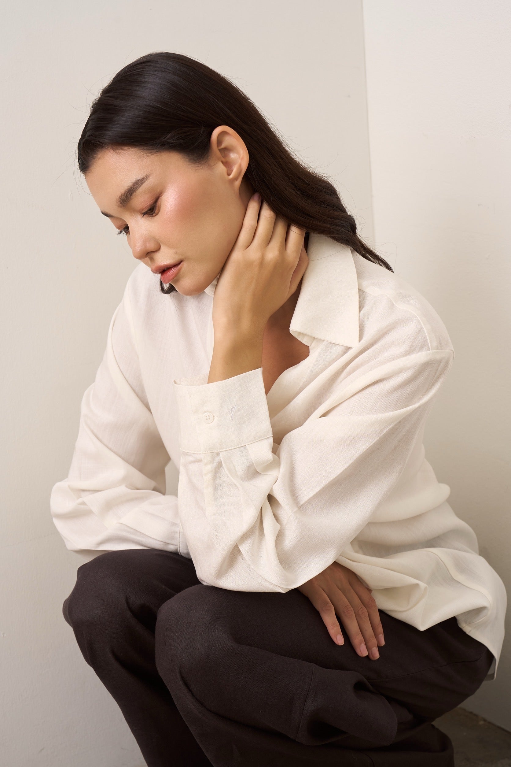 Woman wearing a white linen shirt and dark brown linen pants against a plain background.