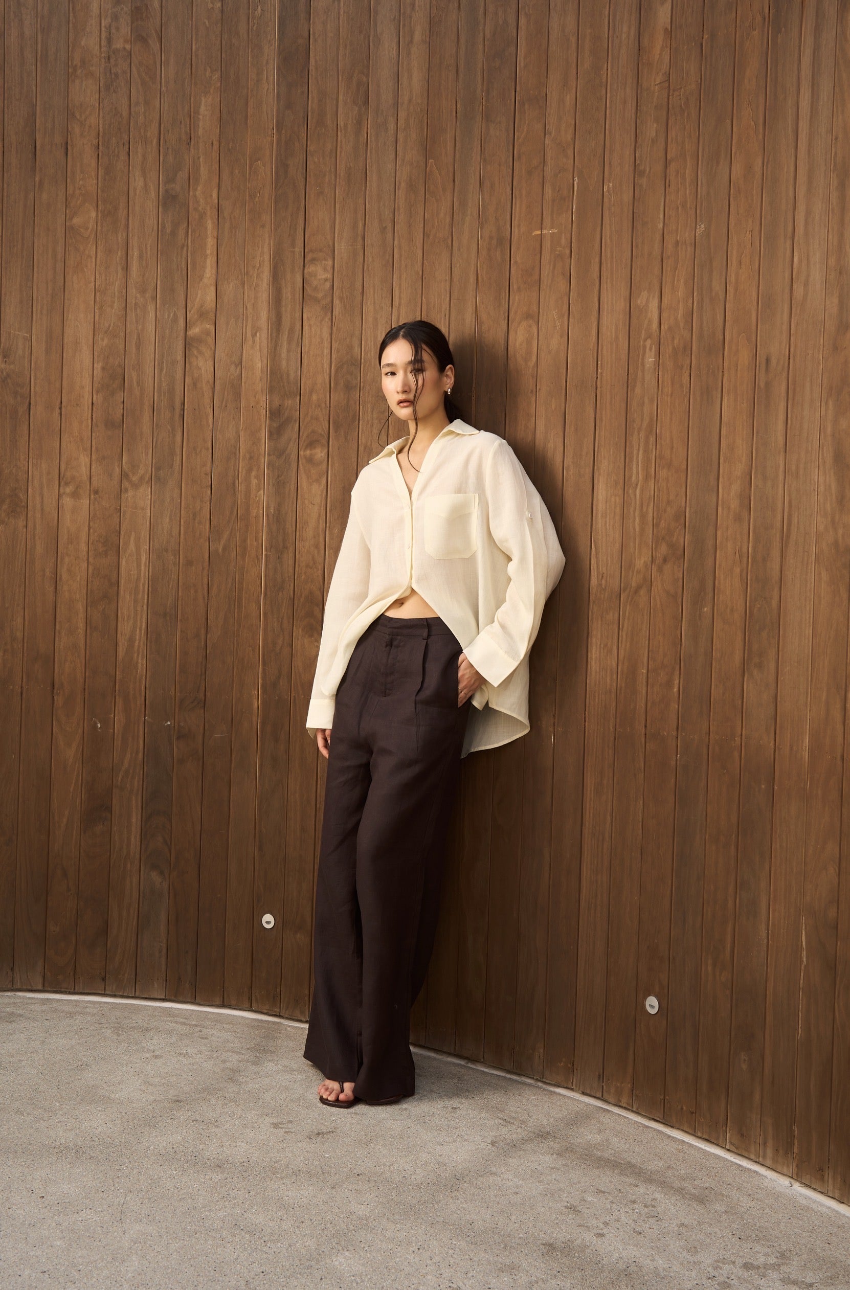 Woman in a butter yellow linen shirt with v collar and dark brown linen pants leaning against a wooden wall.