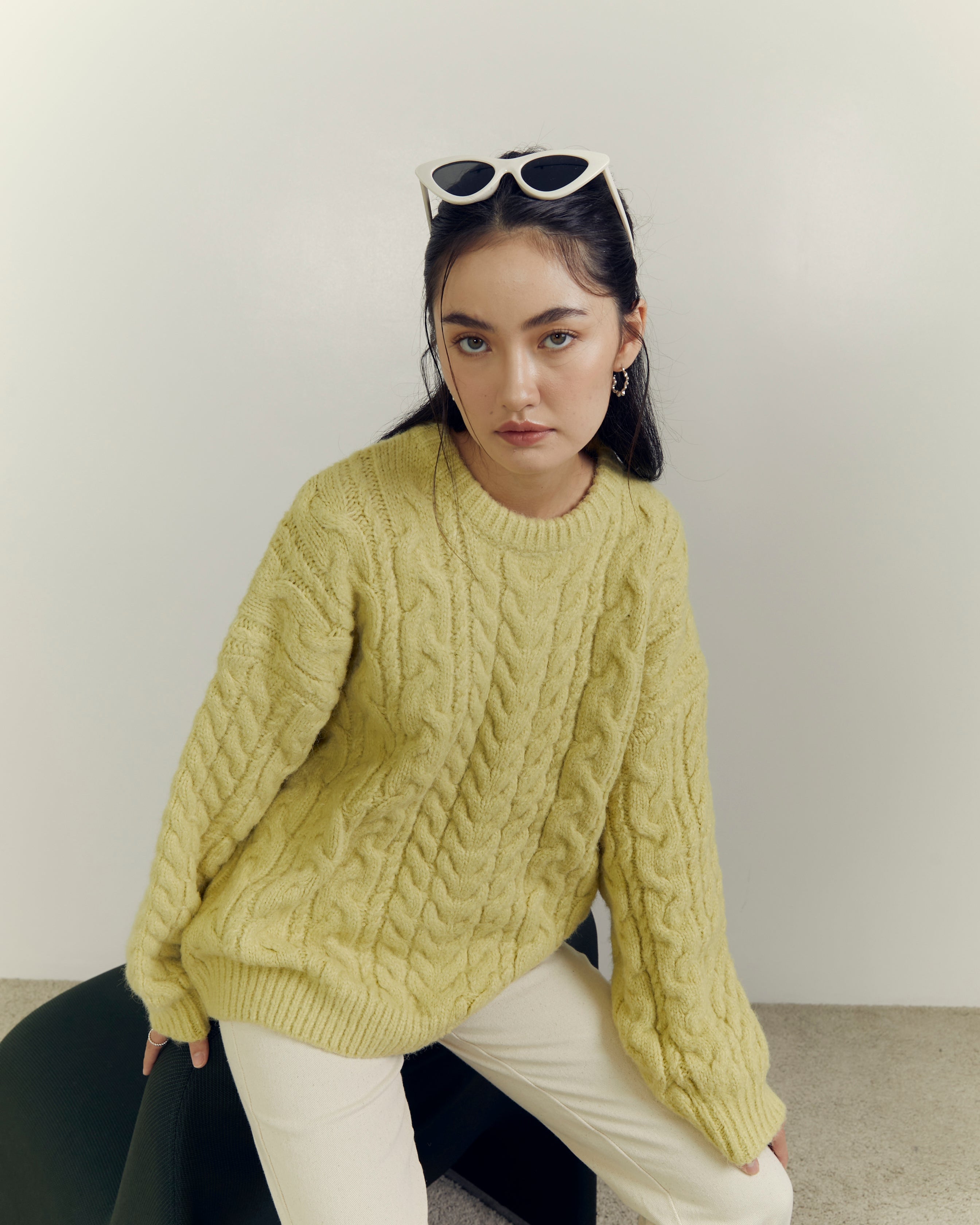 Woman wearing a green cable knit sweater and white pants sitting on a black chair.