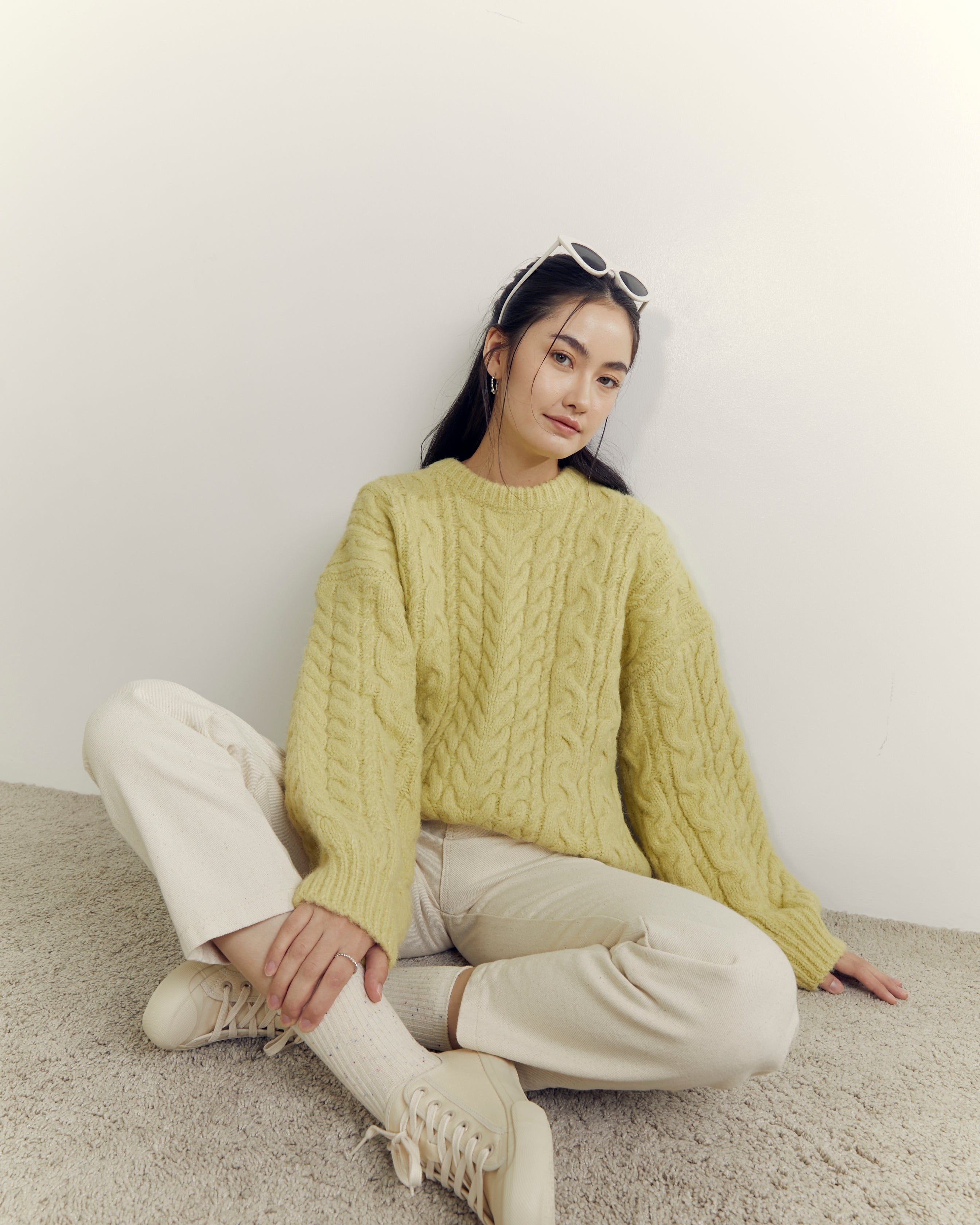Woman wearing a light green cable knit sweater and beige pants sitting on a carpeted floor.