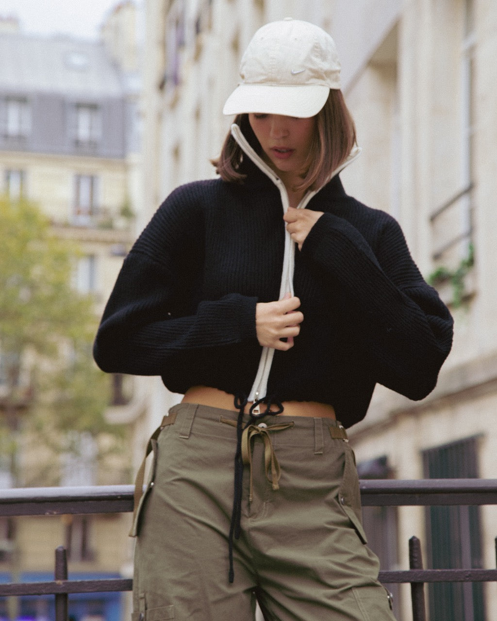 woman wearing a black sweater and beige cargo pants with a white cap in an urban setting.
