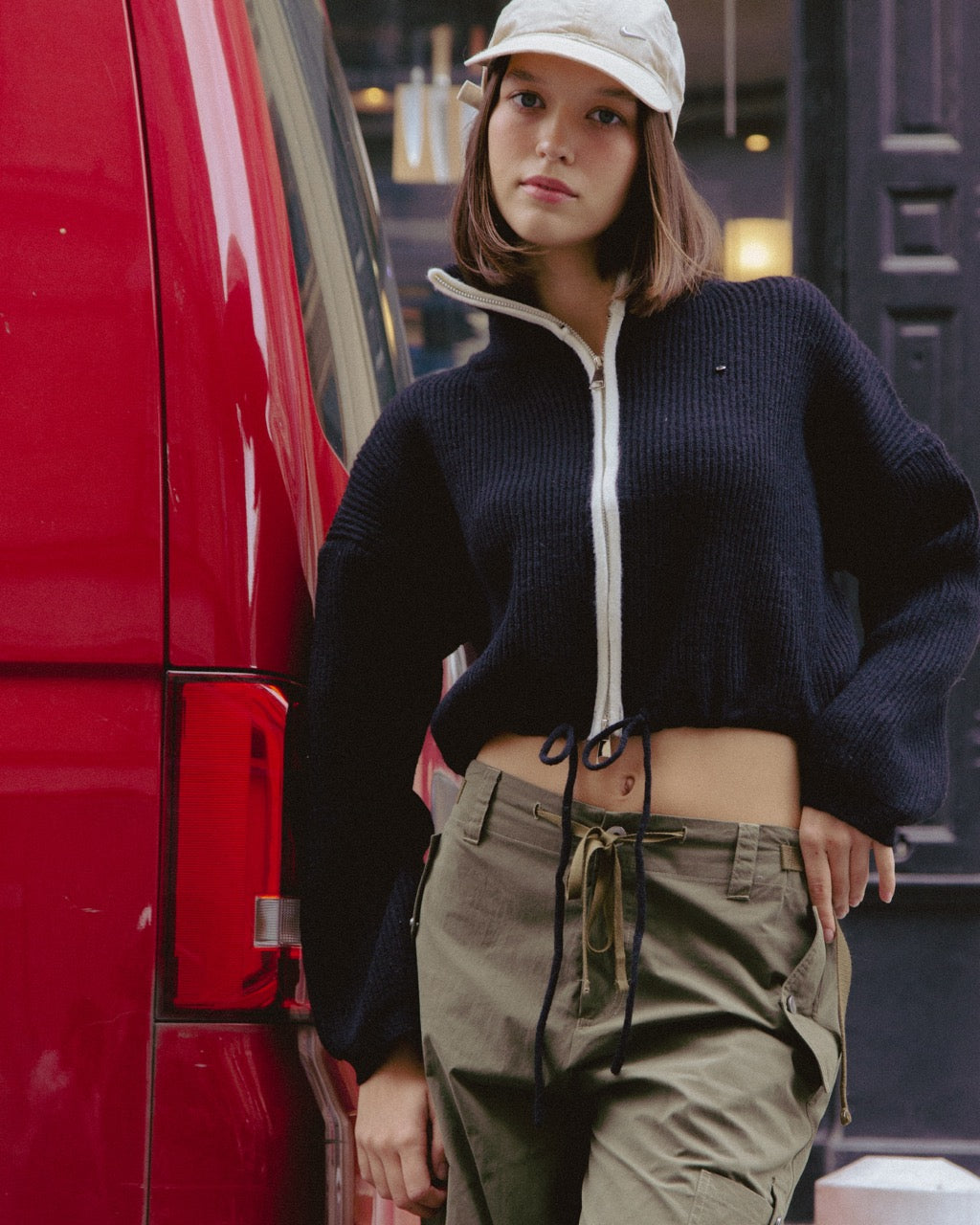 Person wearing a black sweater with a white hood and beige pants standing next to a red vehicle.