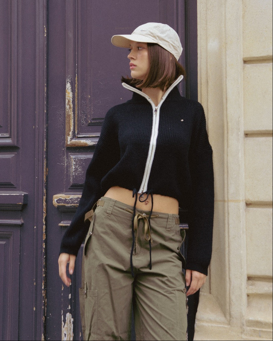 woman wearing a black jacket with a white zipper and beige cap standing in front of a purple door.