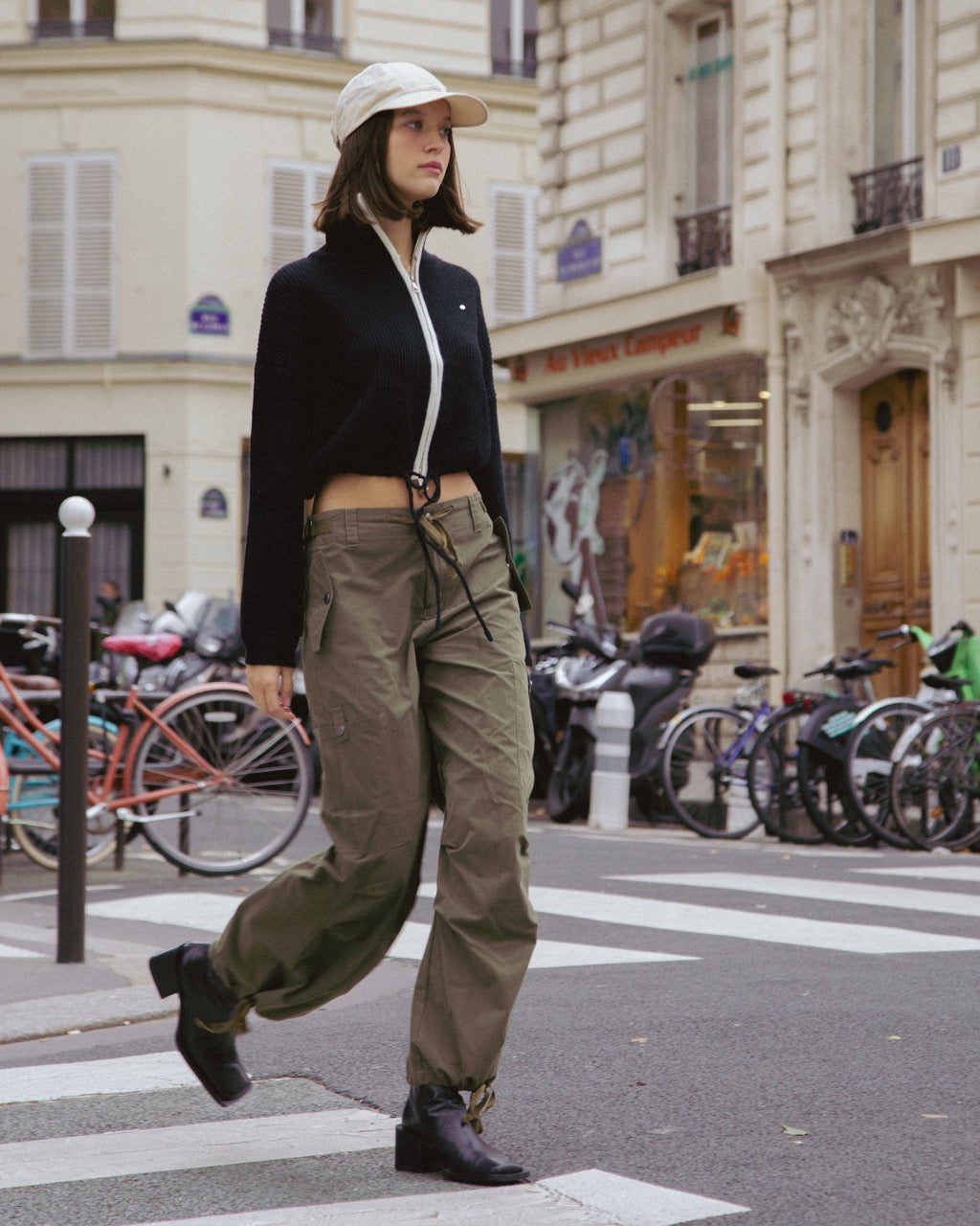 woman walking on a city street wearing a black sweater, beige cap, and green cargo pants.