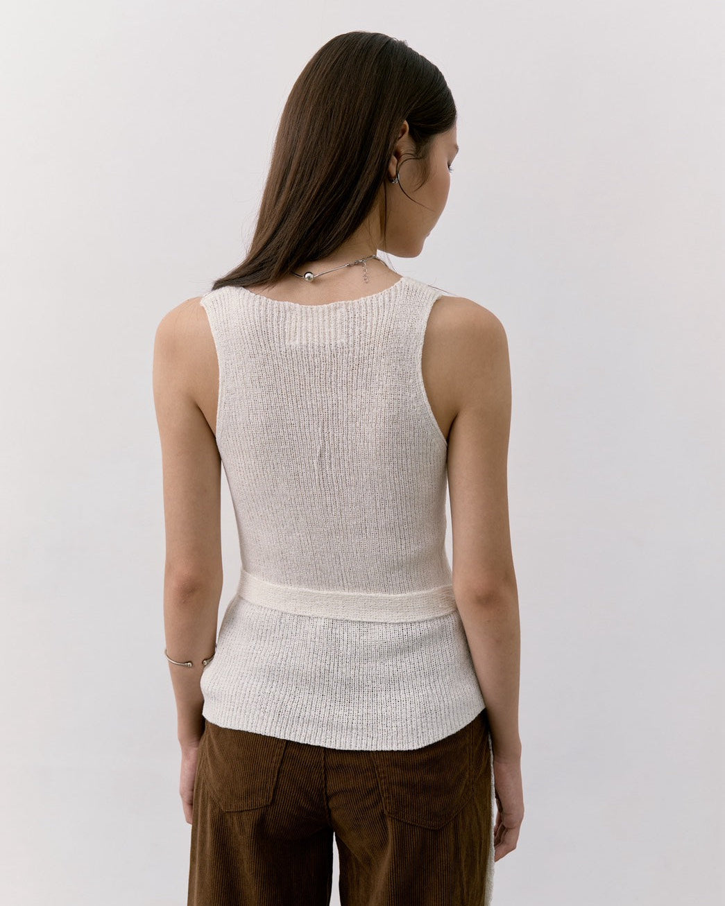 woman viewed from behind wearing a cream ribbed knit sleeveless top and brown corduroy pants against a plain white wall.