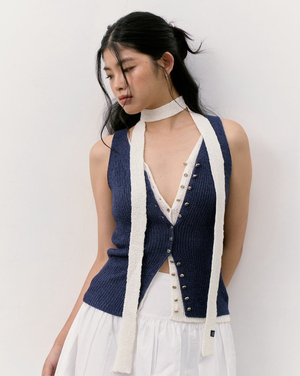 woman wearing a navy blue ribbed knit halter vest with cream-colored trim and buttons, paired with a white high-neck top and white skirt, set against a plain off-white wall.