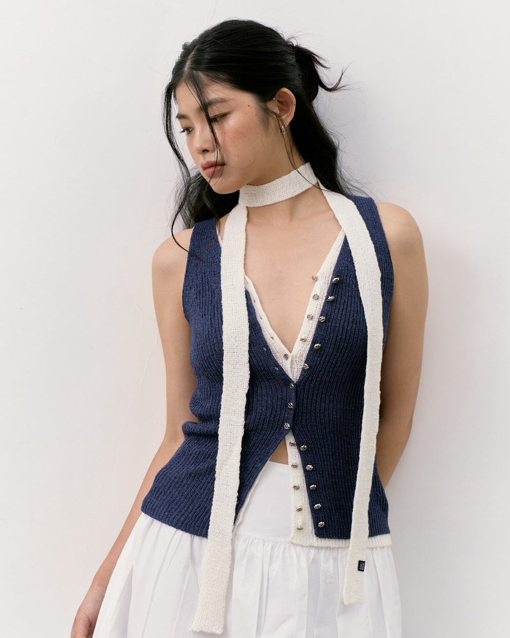 woman wearing a navy blue ribbed knit halter vest with cream-colored trim and buttons, paired with a white high-neck top and white skirt, set against a plain off-white wall.