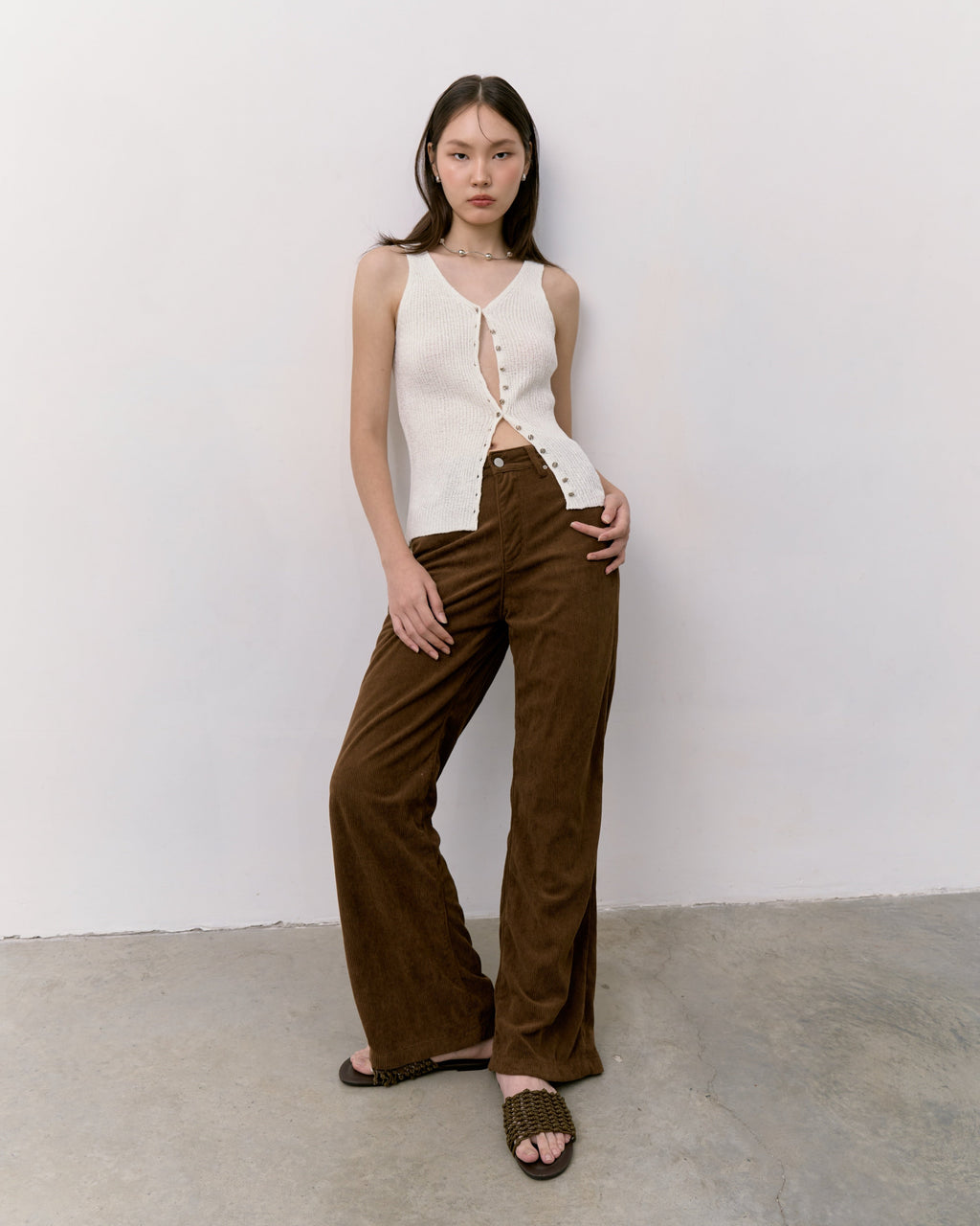 Woman wearing a white sleeveless top and brown pants against a white wall.