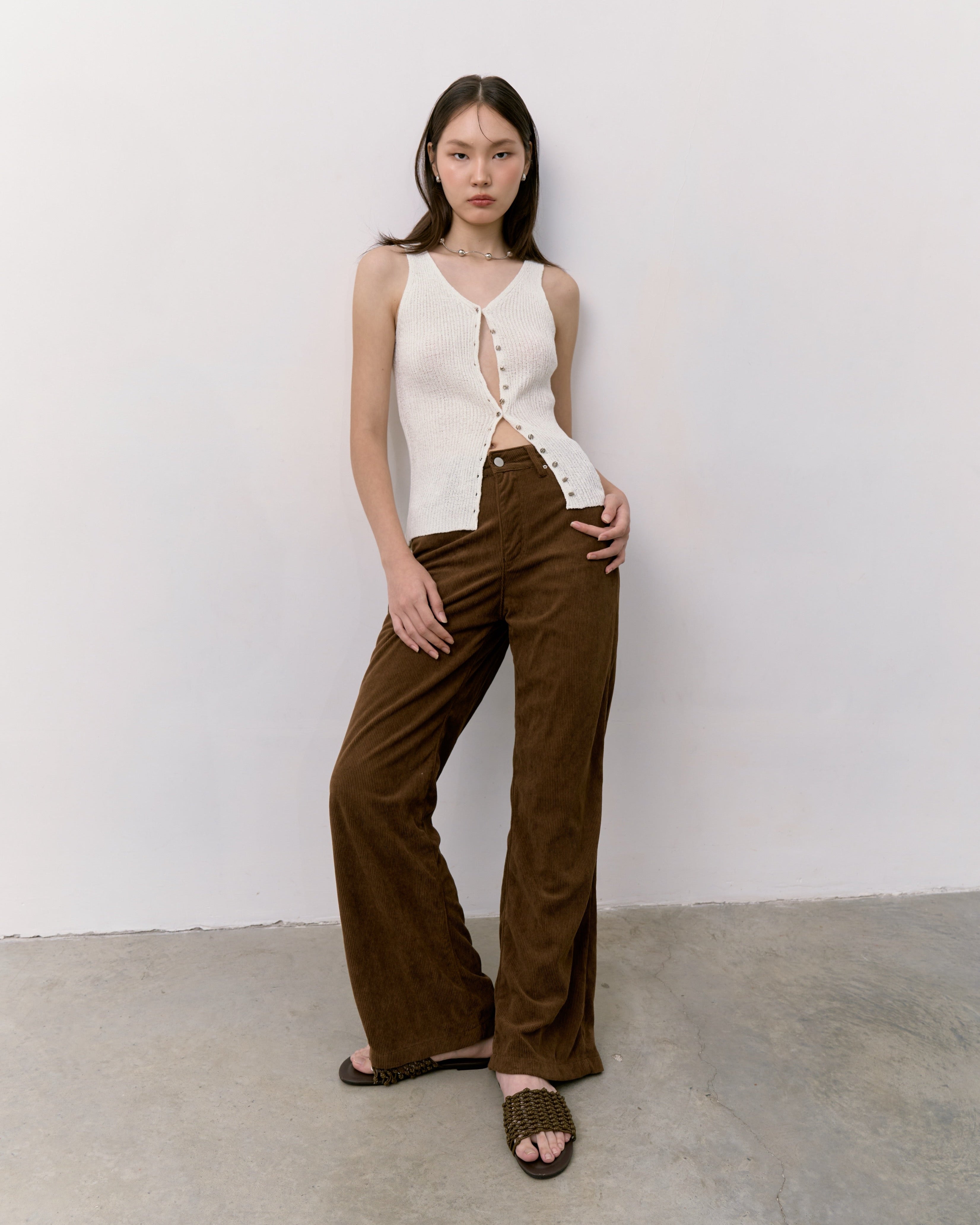 Woman wearing a white sleeveless top and brown pants against a white wall.