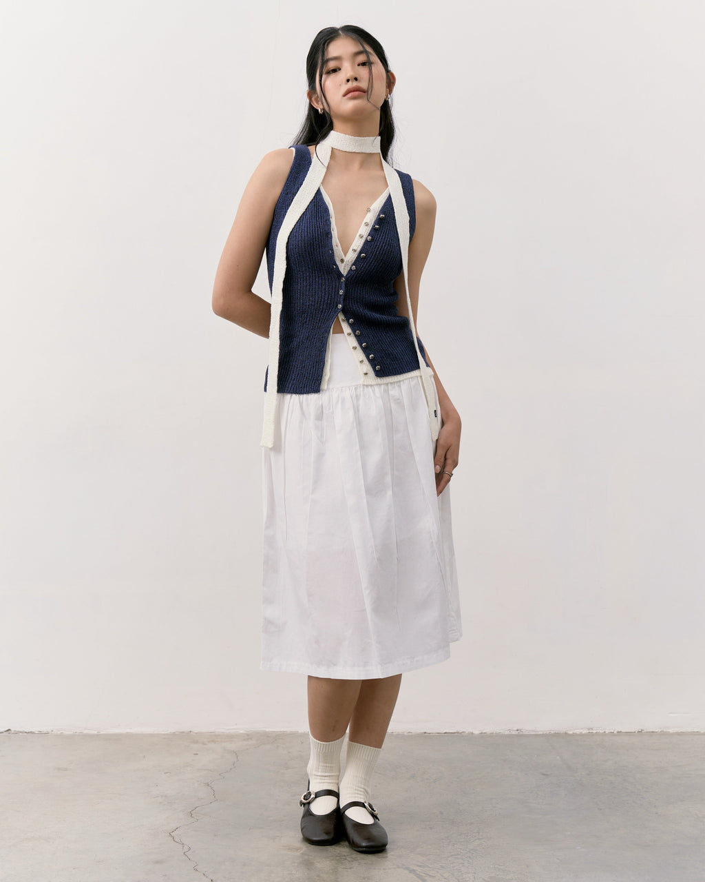 Woman wearing a sleeveless navy top with white trim and a white skirt against a plain background
