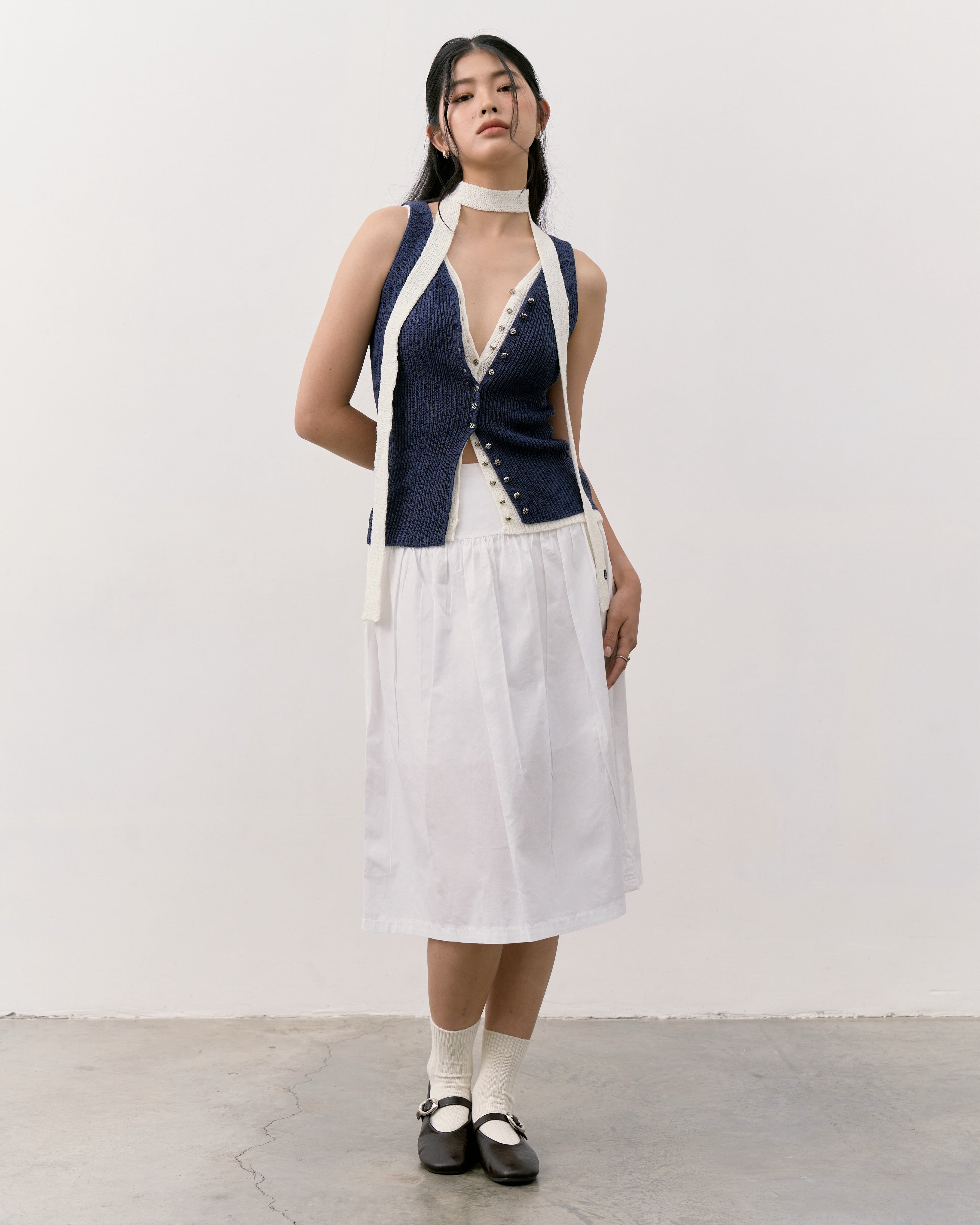 Woman wearing a sleeveless navy top with white trim and a white skirt against a plain background