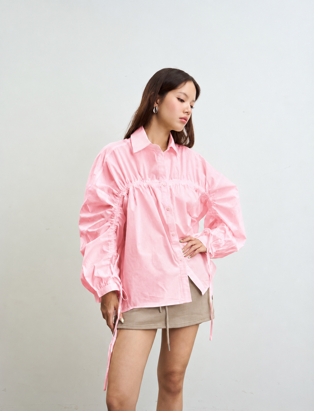 woman modeling a light pink long-sleeve button-up shirt with gathered detailing and drawstring ties, paired with a beige mini skirt, against a plain light gray background.