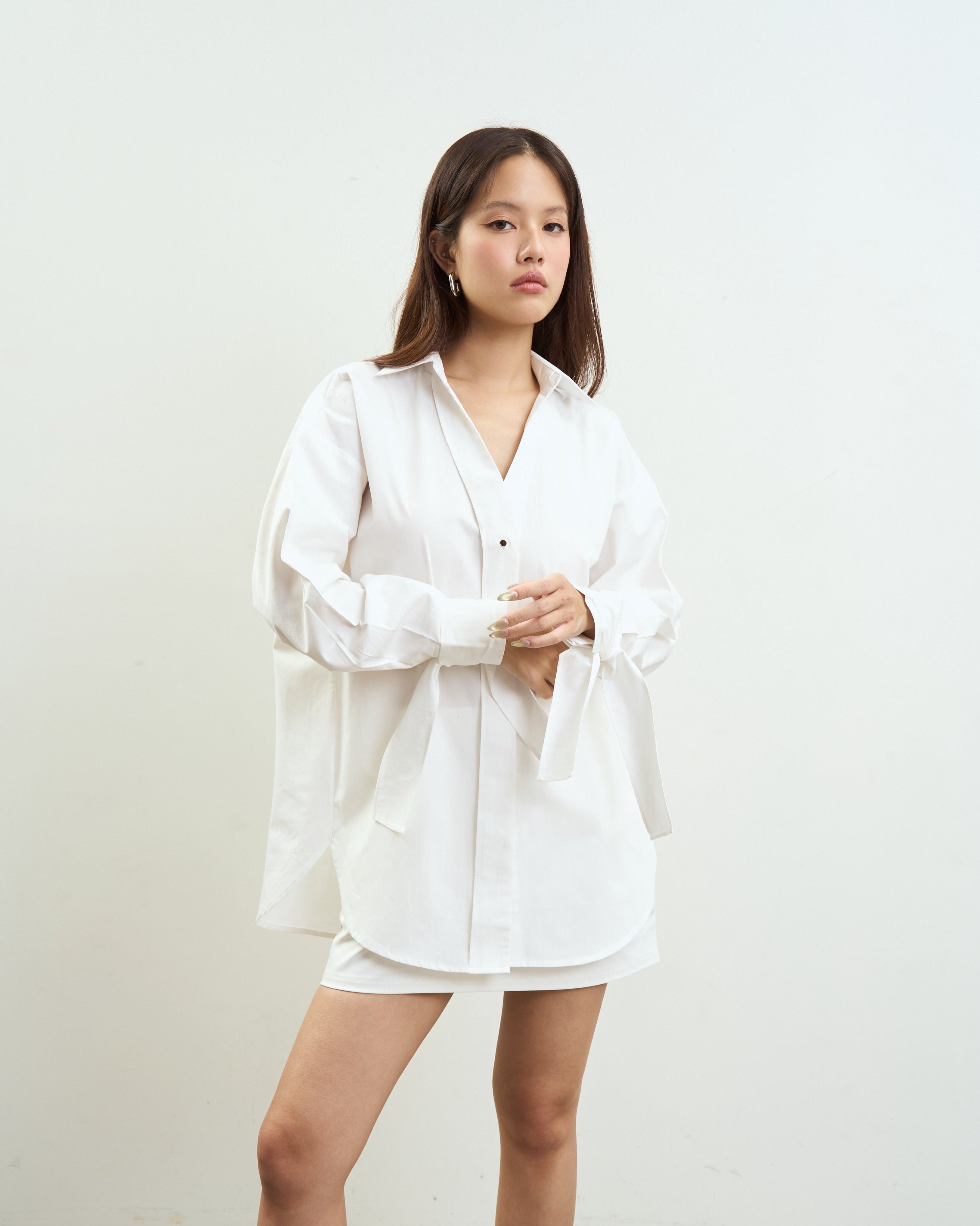 woman modeling a white oversized button-up shirt with matching white shorts against a plain off-white studio wall.