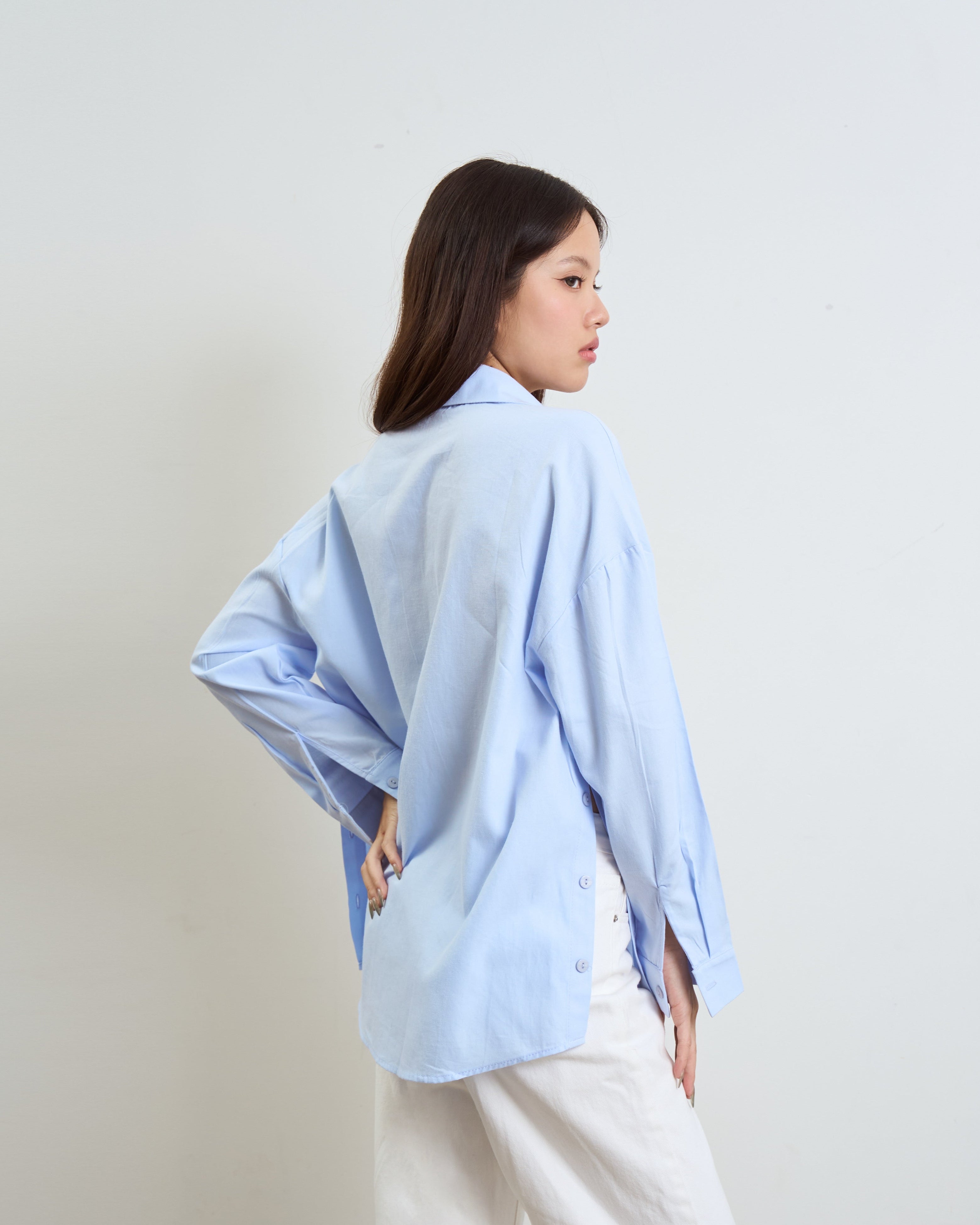 woman wearing an oversized light blue button-up shirt and white pants, photographed from the back against a plain off-white studio background.