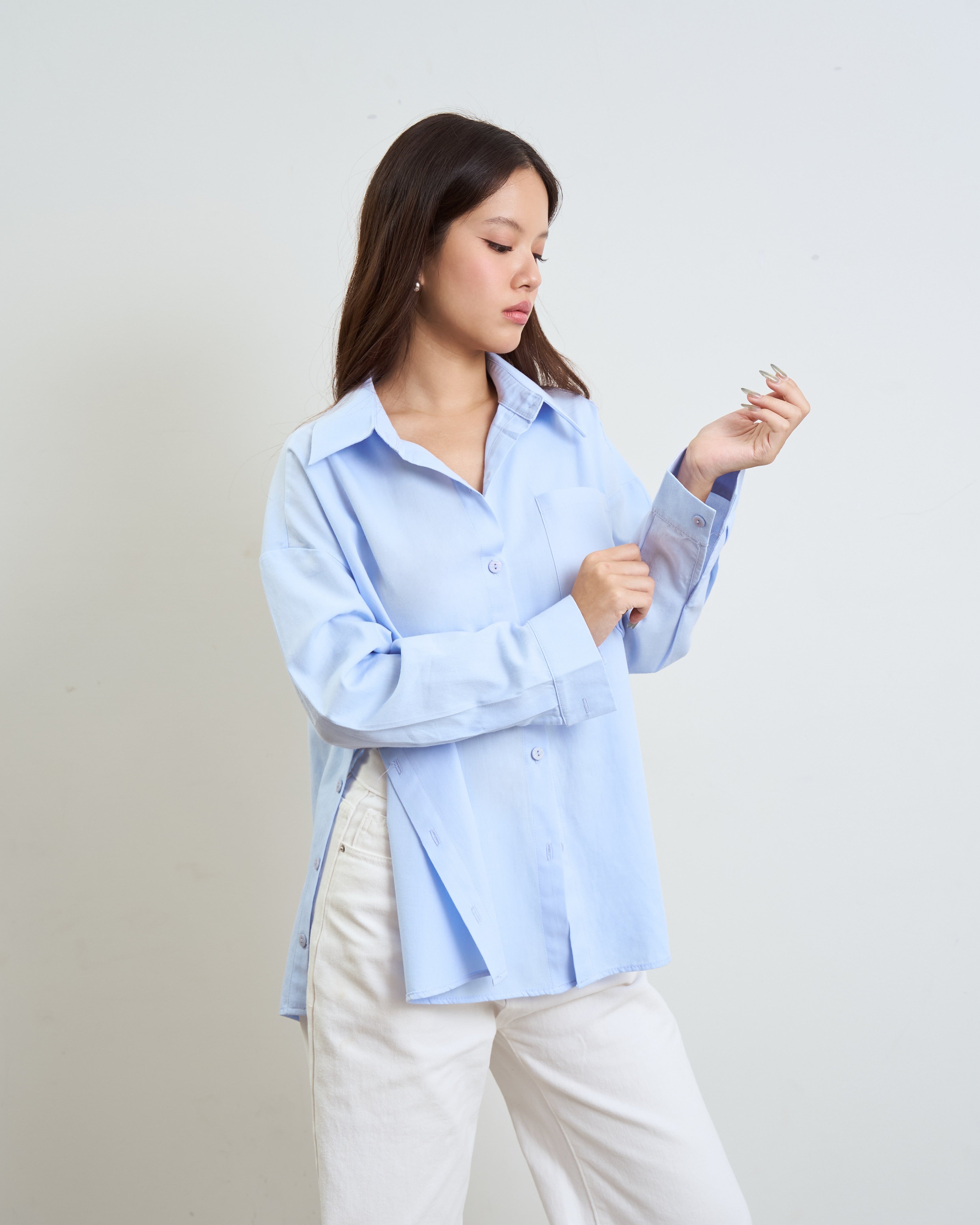 woman wearing a blue button-up shirt and white pants, holding a small silver object in her hand against a plain light gray background.