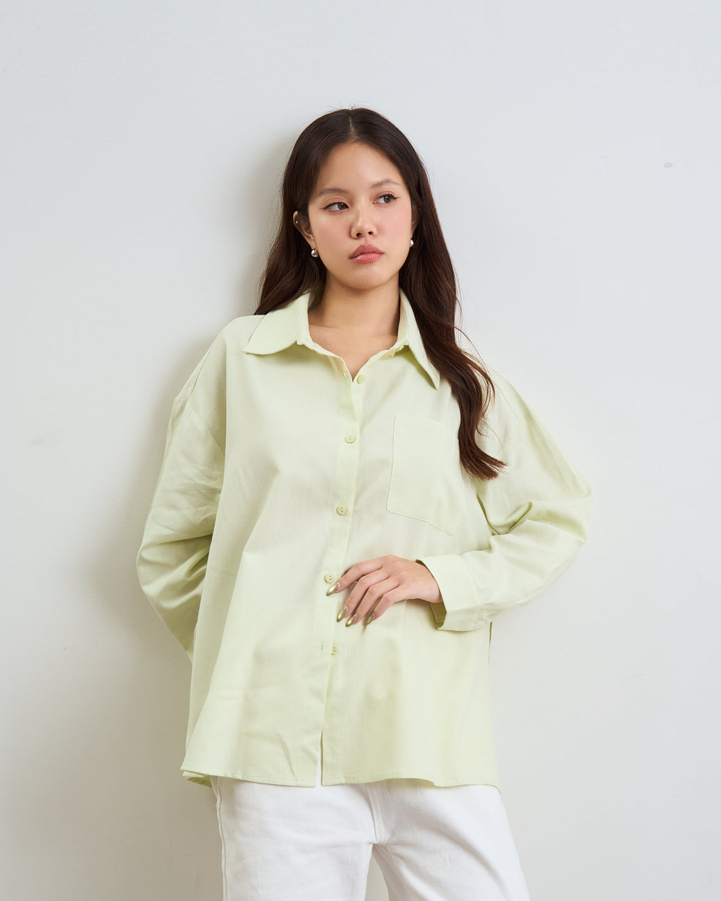 woman wearing a pale yellow-green long-sleeve button-up shirt and white pants, standing against a plain light gray wall.