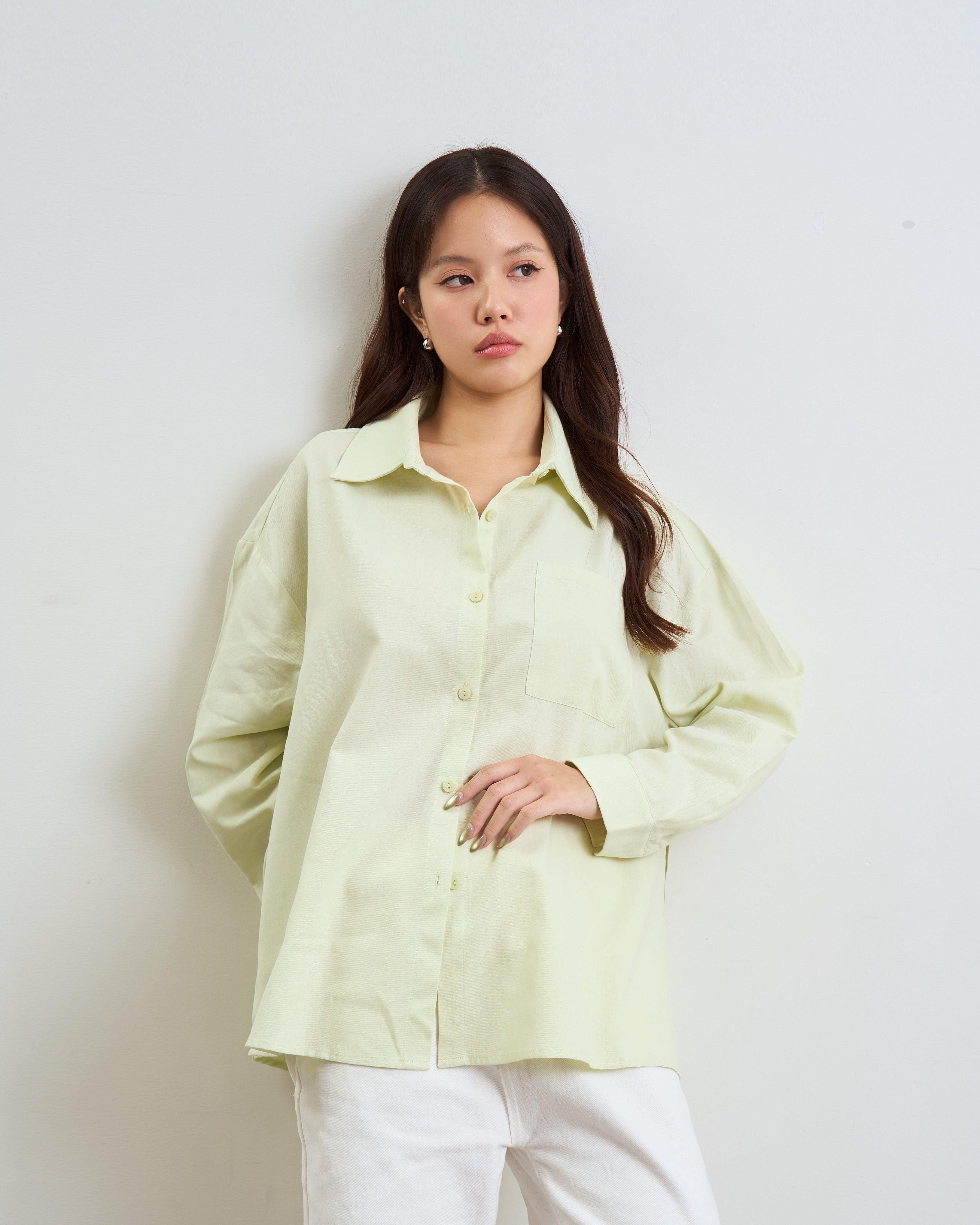 woman wearing a pale yellow-green long-sleeve button-up shirt and white pants, standing against a plain light gray wall.