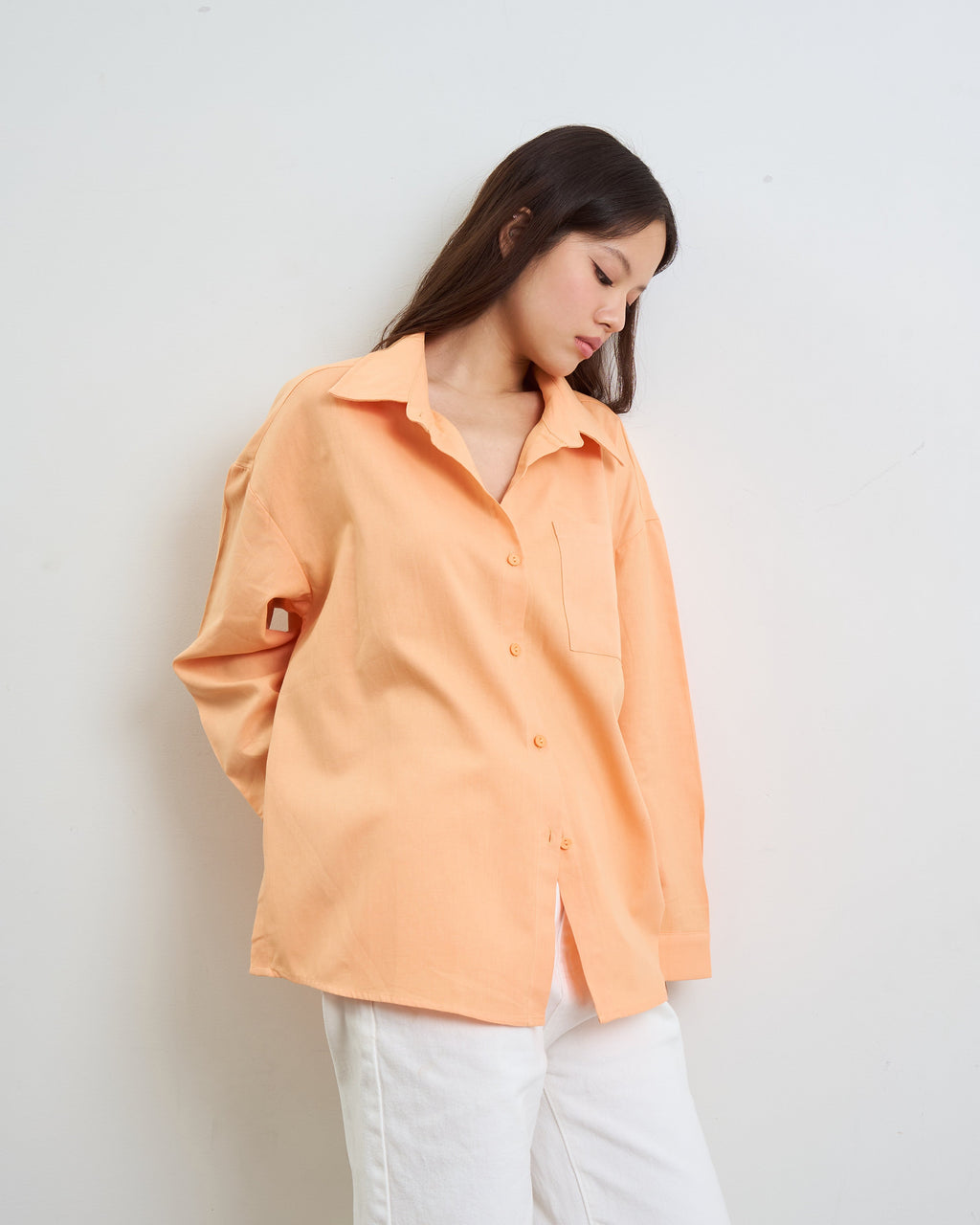 woman wearing a light peach-orange button-up shirt and white pants, standing against a plain light gray wall.