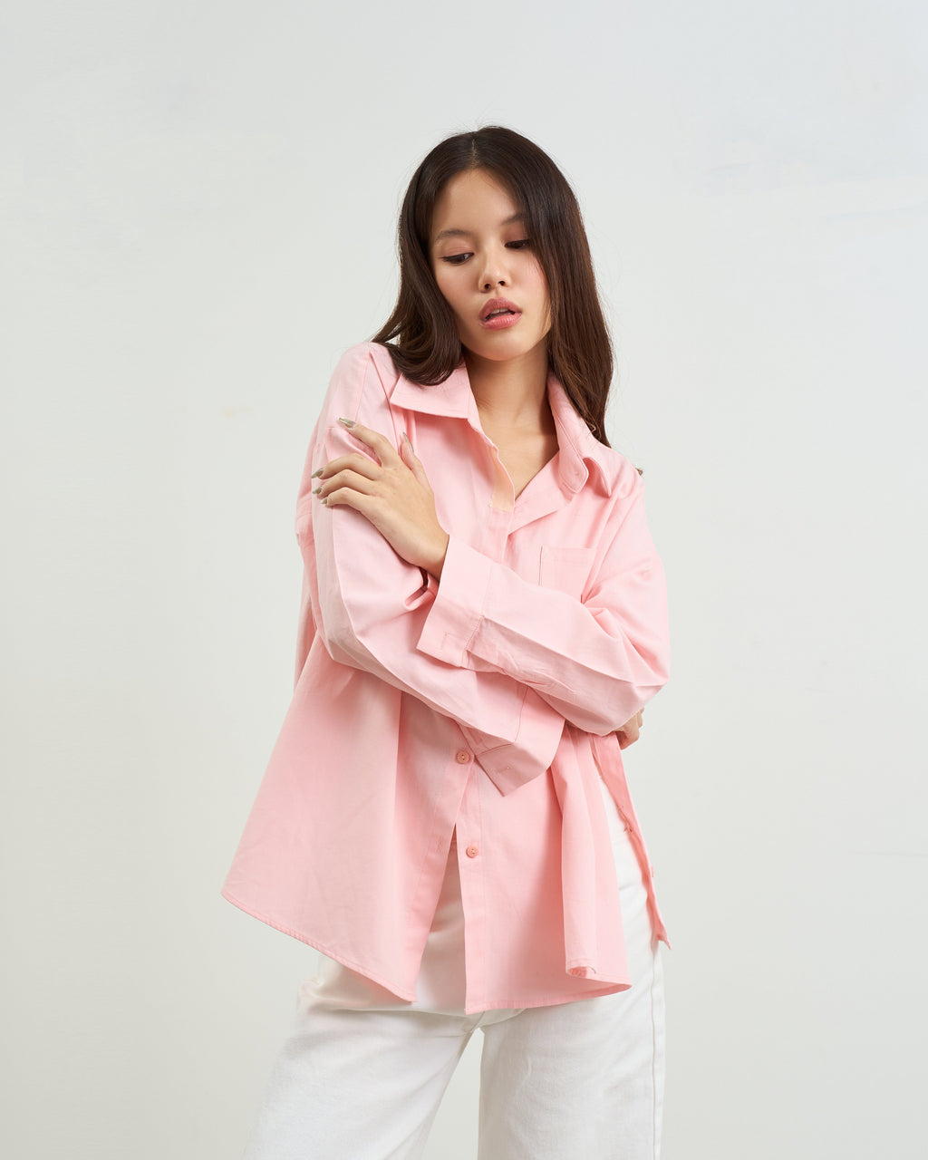 woman modeling a pink oversized button-up shirt with white pants, standing against a plain light gray studio background.