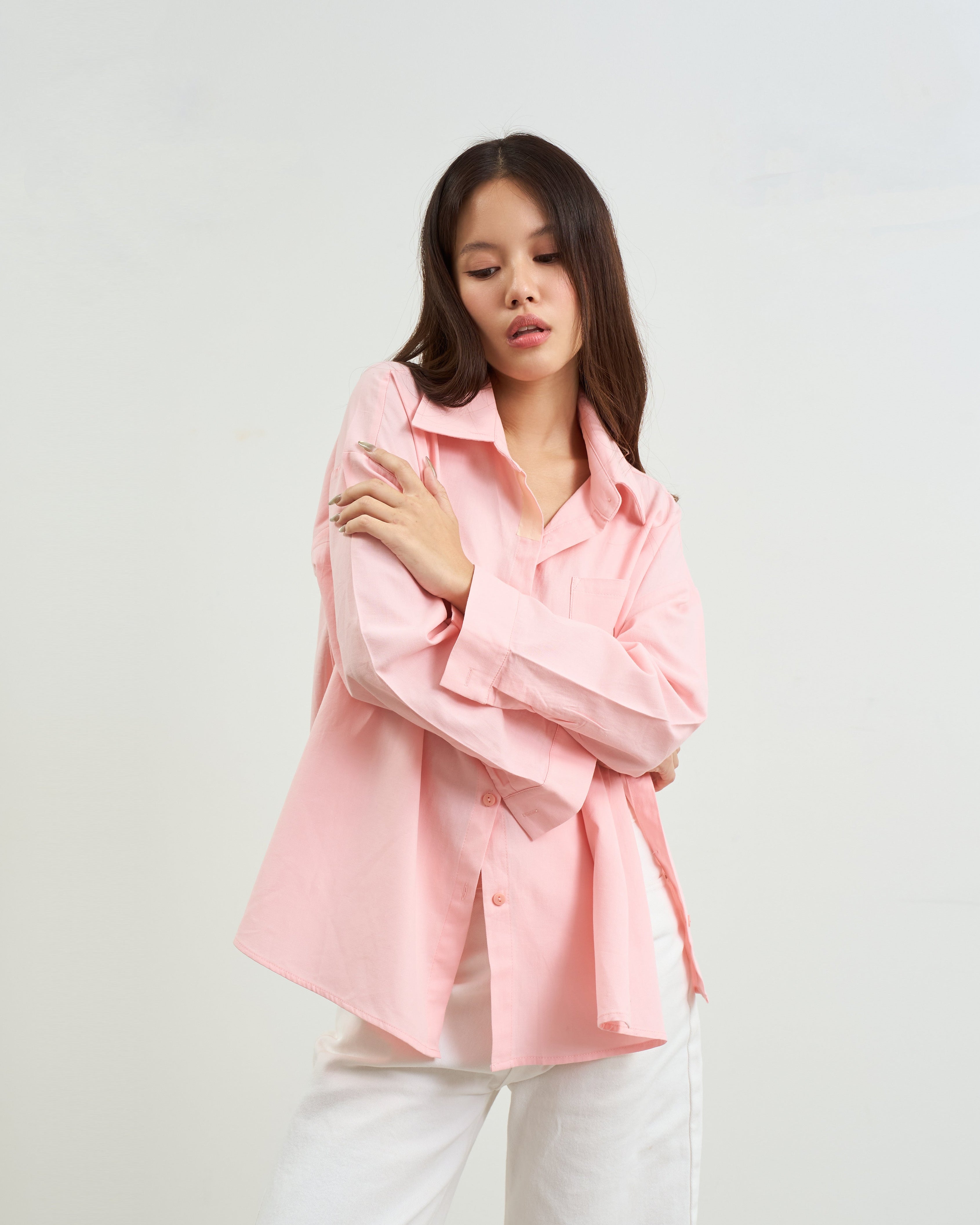 woman modeling a pink oversized button-up shirt with white pants, standing against a plain light gray studio background.