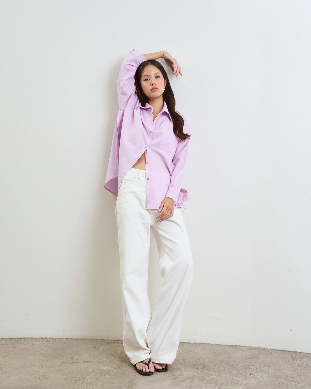 woman posing in a studio setting wearing a purple long-sleeve button-up shirt, white wide-leg trousers, and black sandals against a plain off-white wall and concrete floor.