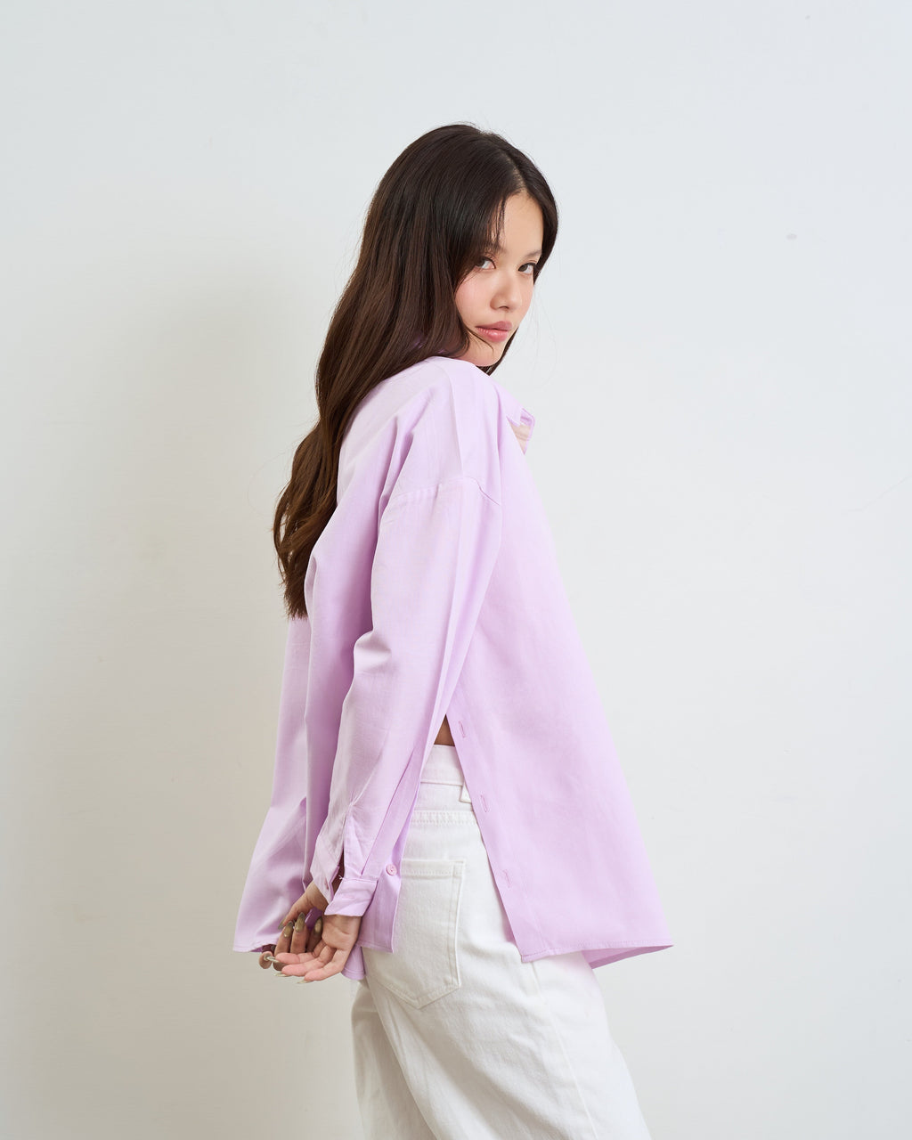 woman wearing a purple long-sleeve shirt with side slits and white pants, photographed against a plain light gray studio background.