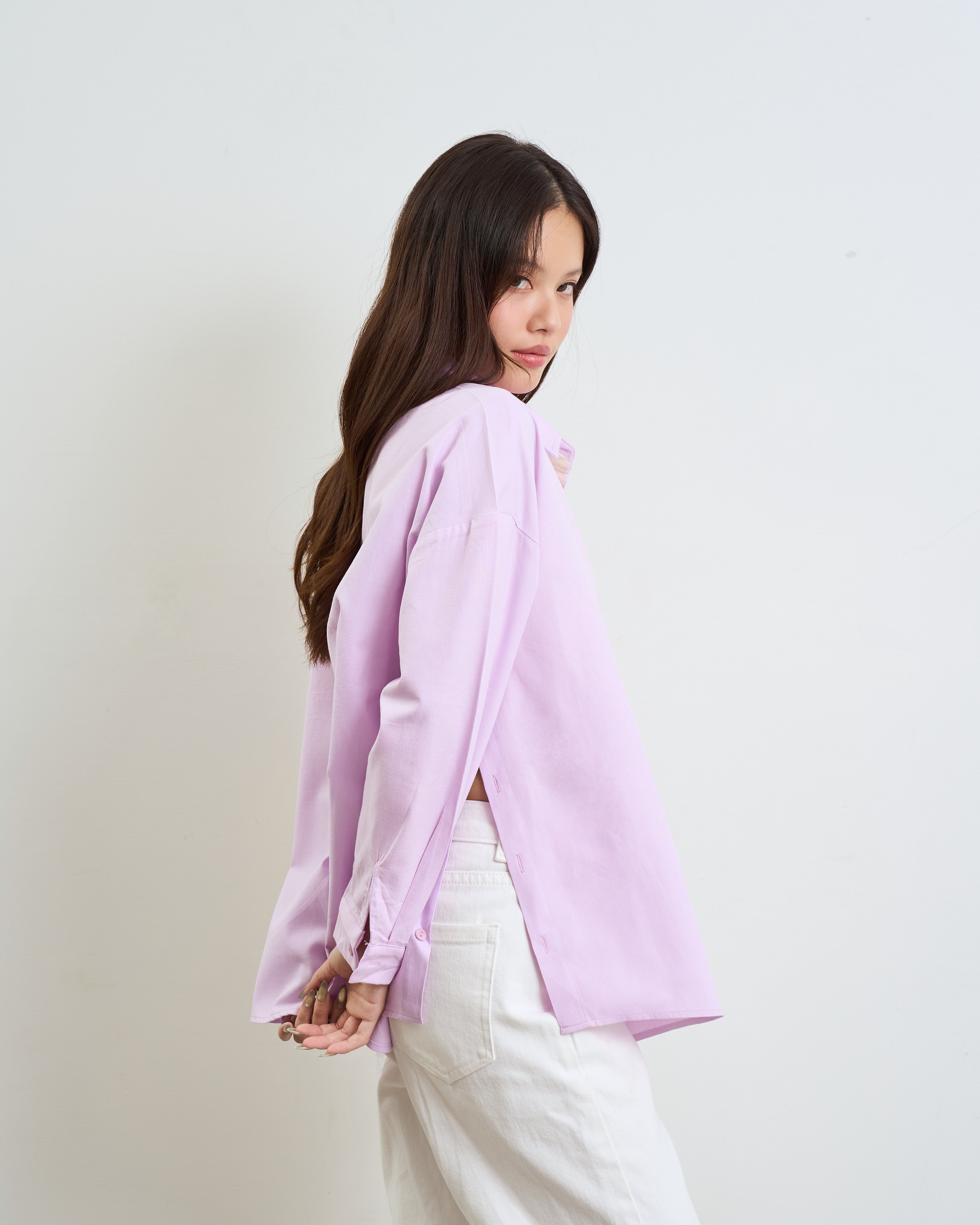 woman wearing a purple long-sleeve shirt with side slits and white pants, photographed against a plain light gray studio background.