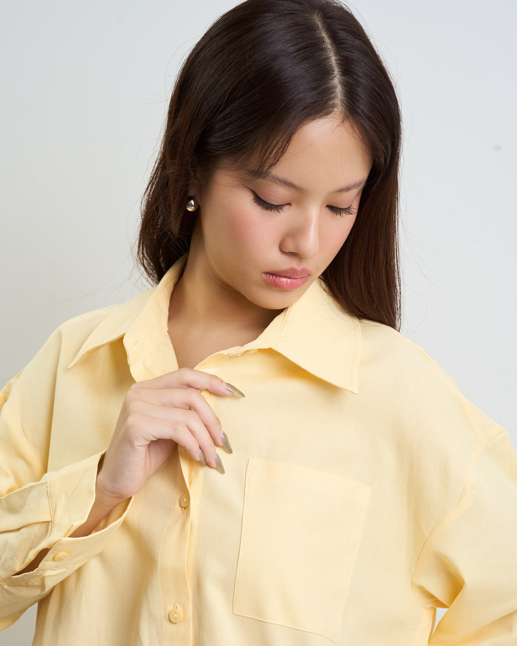 woman wearing an oversized pale yellow button-up shirt with a chest pocket, shown from the shoulders down against a light gray studio background.