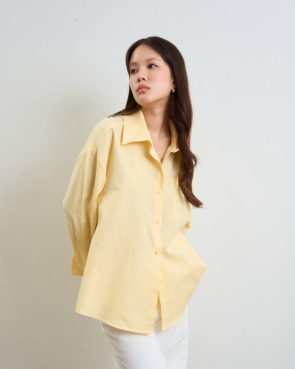 woman wearing a pale yellow oversized button-up shirt and white pants, standing against a plain light gray wall.