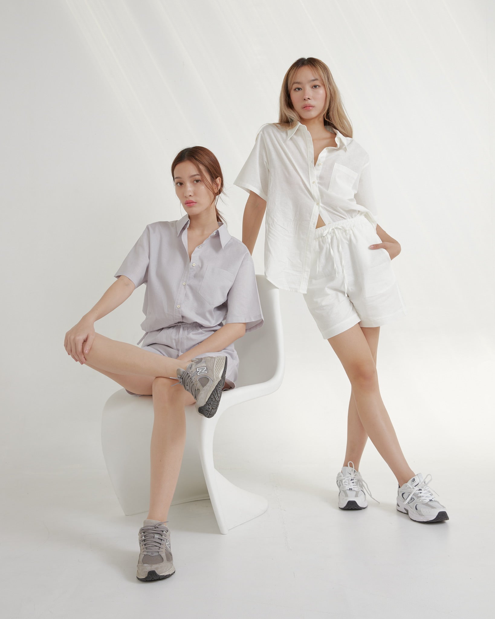 Two women in light-colored outfits sitting on a white chair against a white background