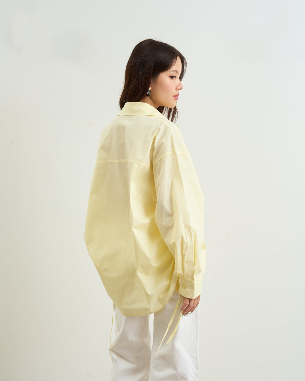 woman shown from the back wearing a loose, oversized yellow button-up shirt and white wide-leg pants, standing against a plain off-white wall.