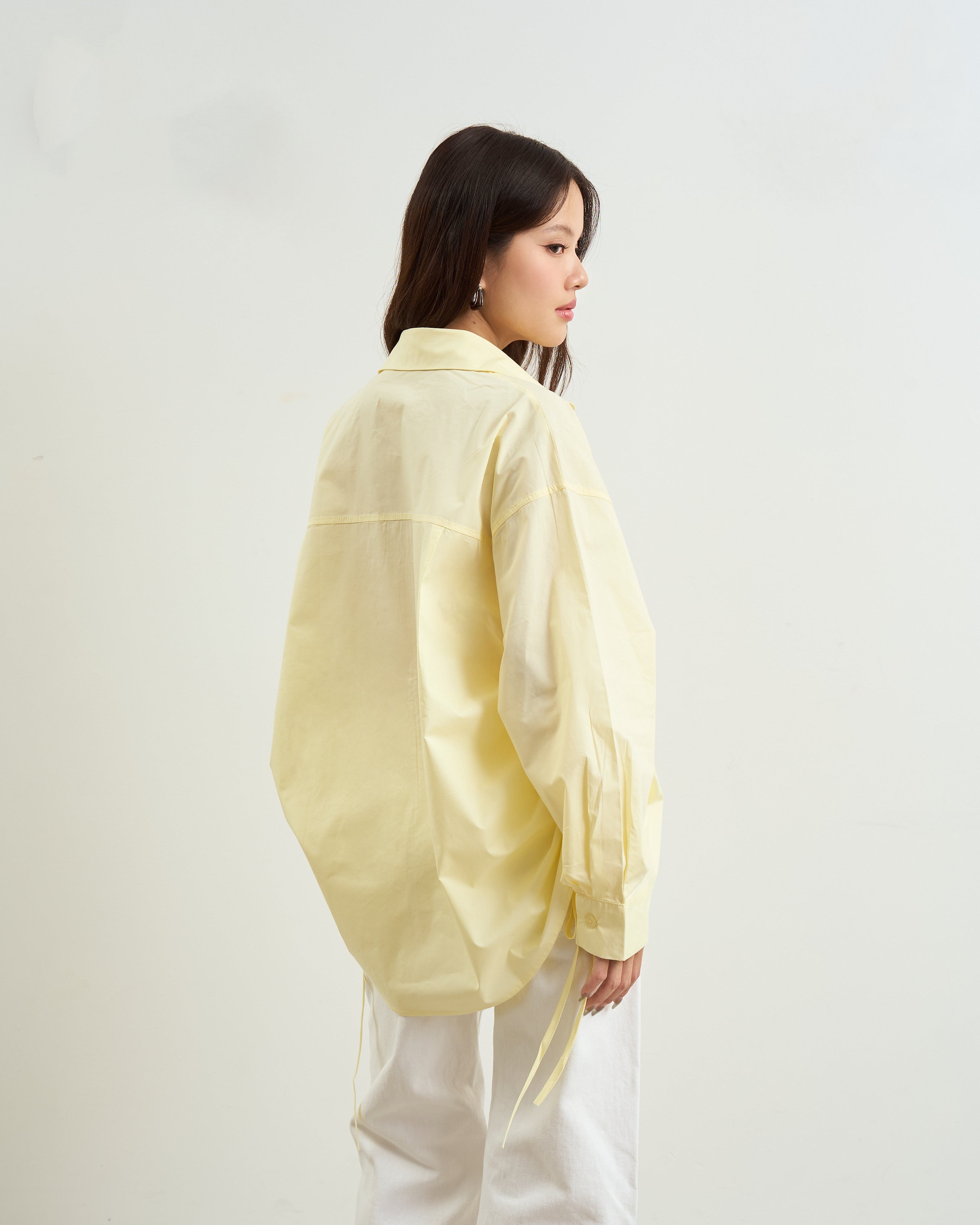 woman shown from the back wearing a loose, oversized yellow button-up shirt and white wide-leg pants, standing against a plain off-white wall.