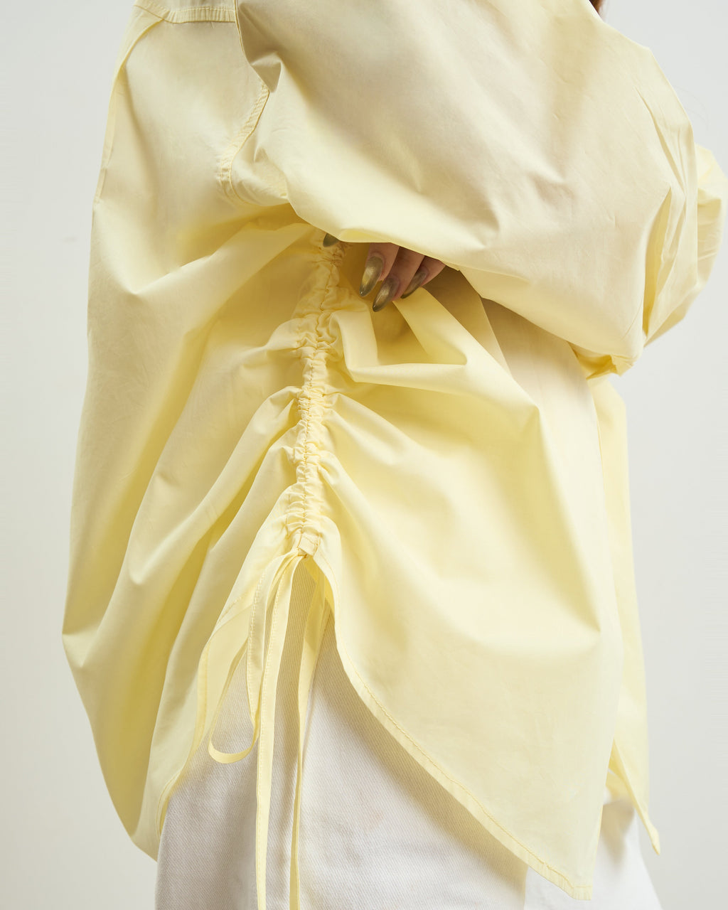 A close-up of a woman wearing a loose, oversized yellow top with a gathered side drawstring and white pants against a plain light background.