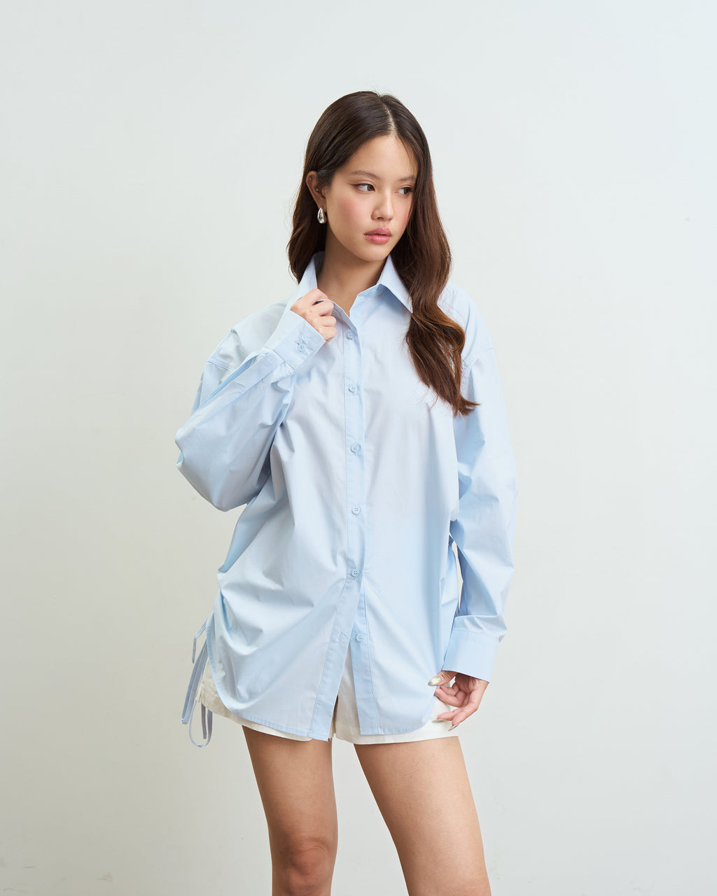 woman modeling an oversized blue button-up shirt with white shorts, standing against a plain off-white wall.