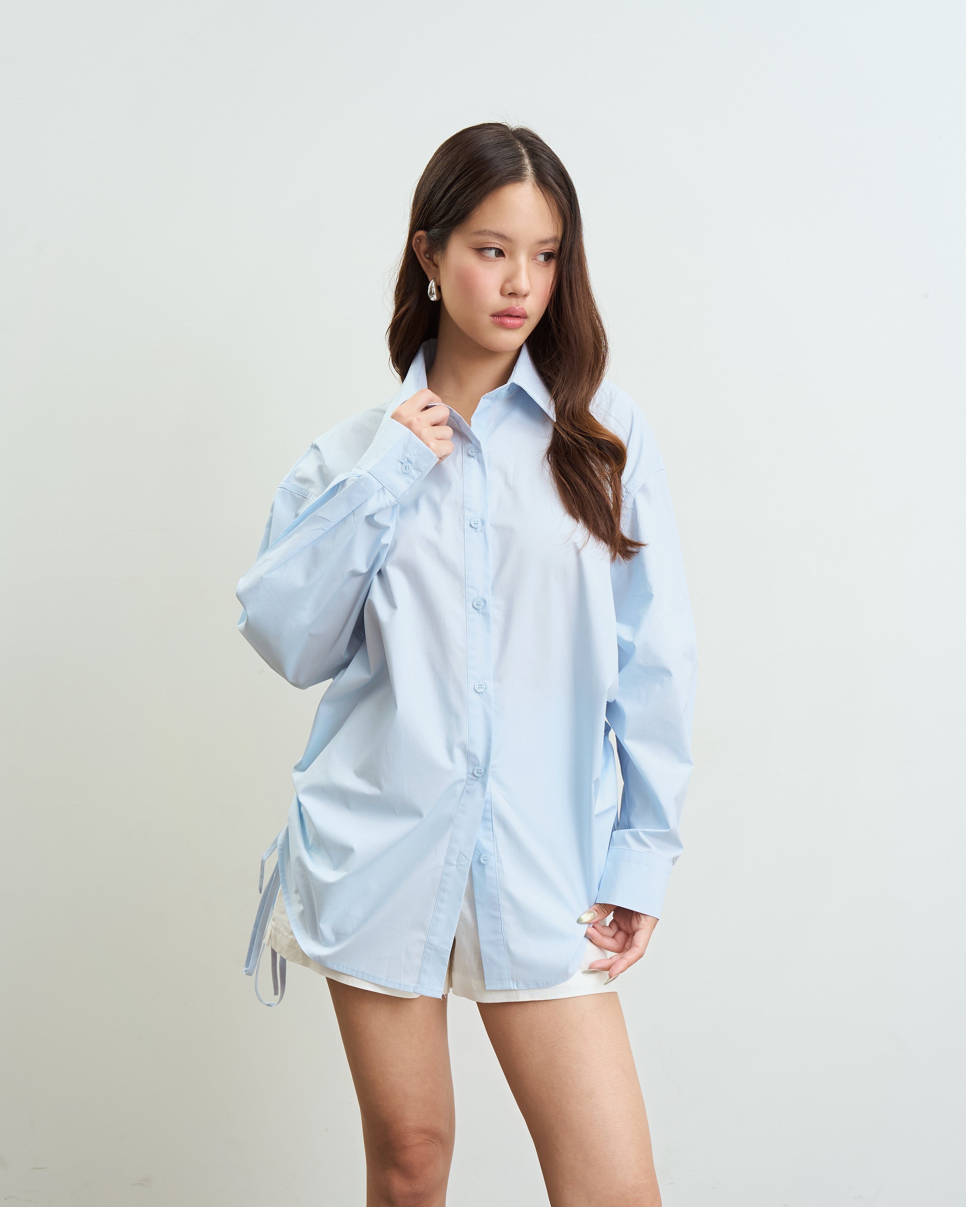 woman modeling an oversized blue button-up shirt with white shorts, standing against a plain off-white wall.