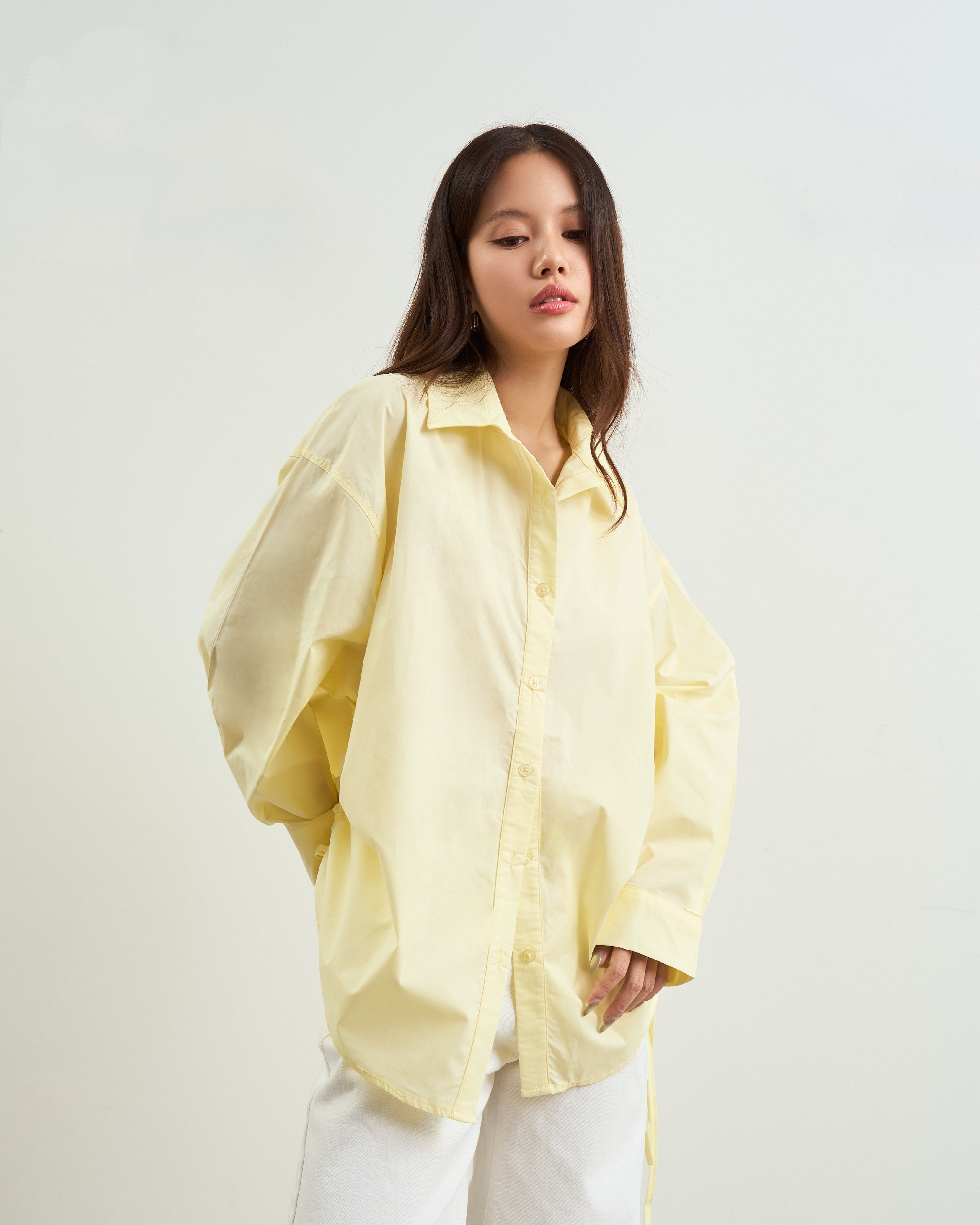 woman wearing an oversized yellow button-up shirt and white pants, standing against a plain light gray studio background.