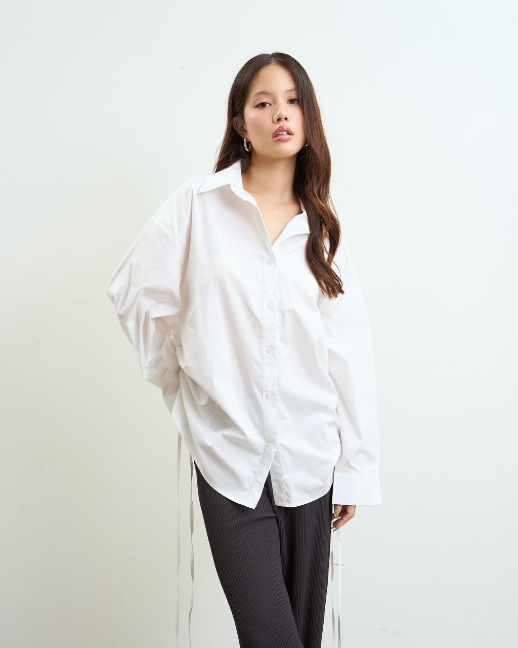 woman modeling an oversized white button-up shirt with dark gray pants, standing against a plain off-white studio background.